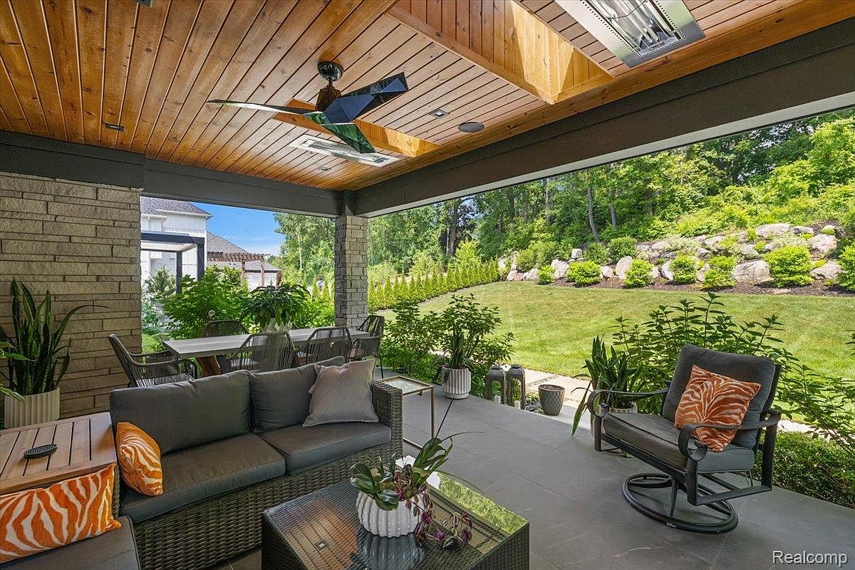 This image showcases a luxurious outdoor patio area, featuring comfortable seating arrangements including a wicker sofa and swivel chair with decorative pillows. The space is covered by a wooden ceiling with a ceiling fan and recessed lighting, offering both shade and ambiance. Lush landscaping surrounds the patio, providing a serene and private outdoor living experience.