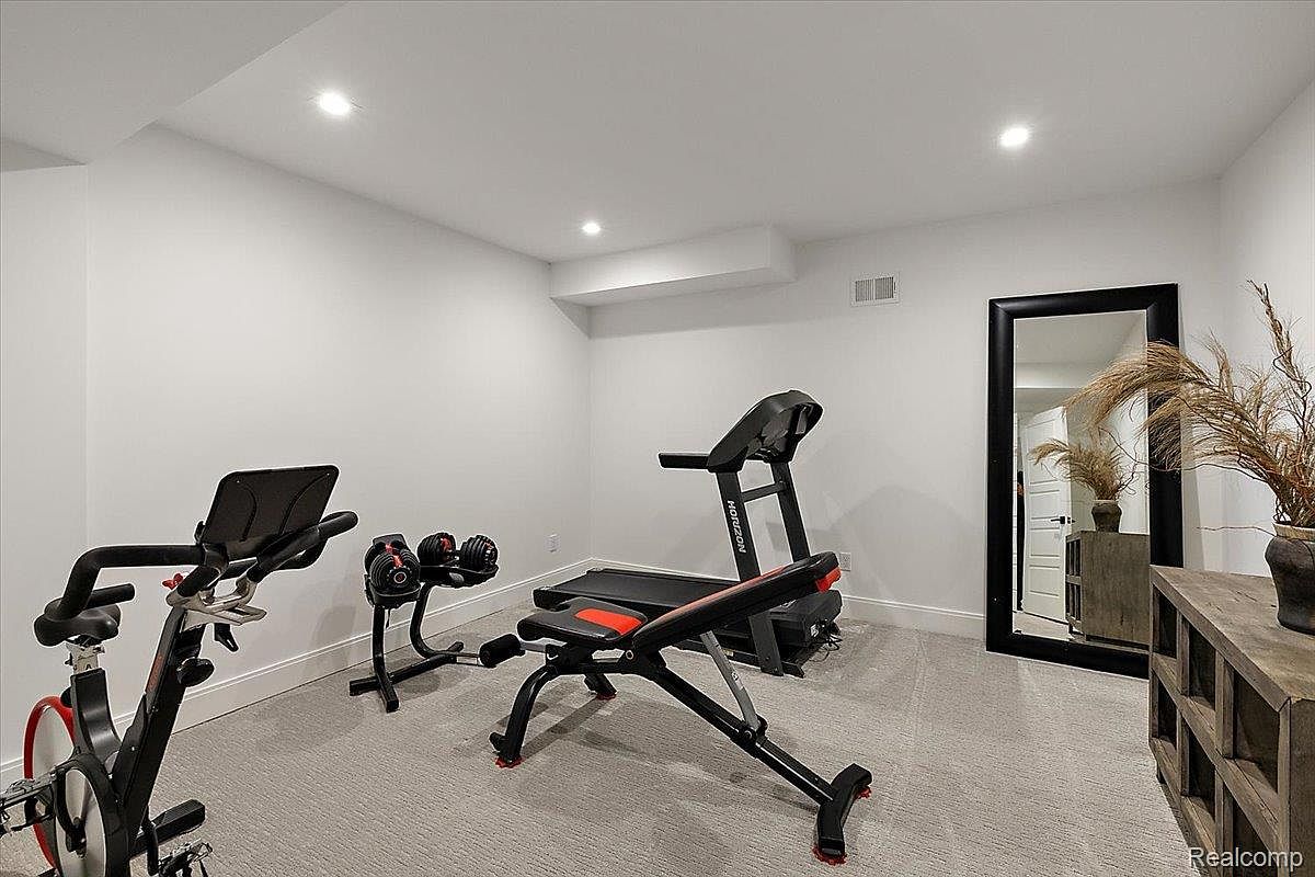 This is a well-lit home gym featuring a stationary bike, adjustable dumbbells, a weight bench, and a treadmill. The walls are painted white, and the floor is covered with gray carpet. A large mirror leans against the wall, adding depth to the space, and a wooden console table with decorative plants sits to the right.