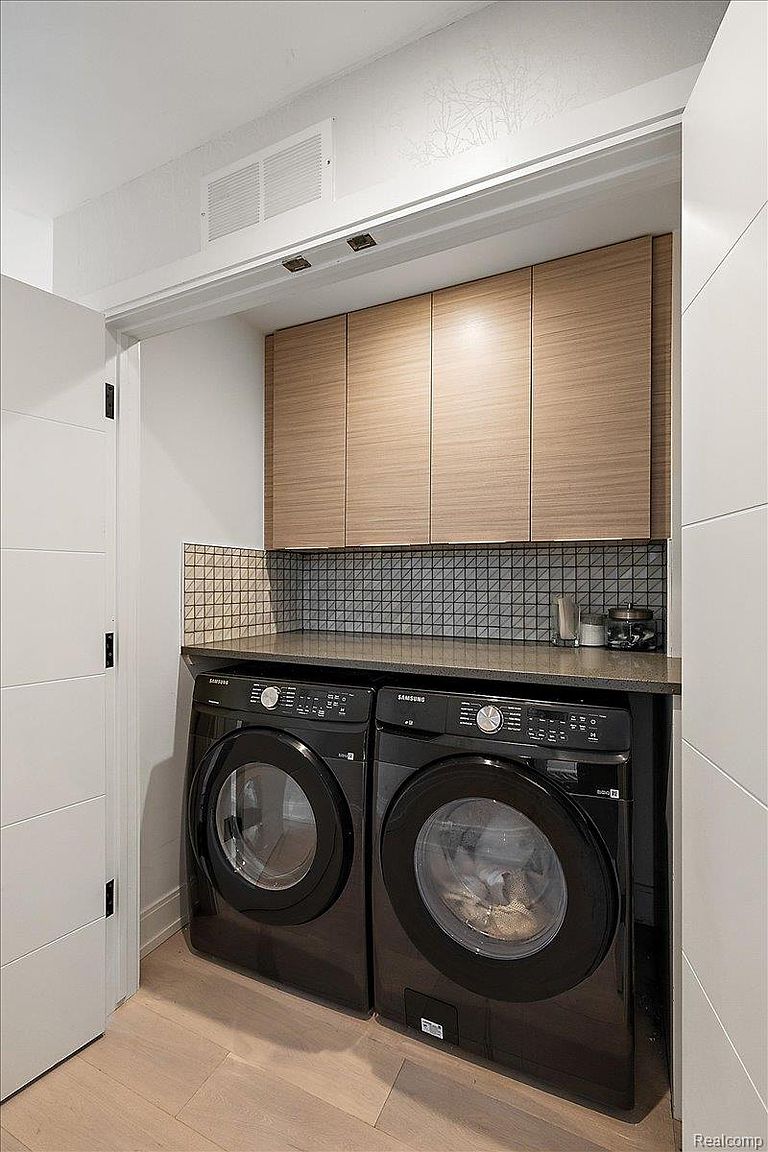 This is a well-organized laundry room featuring a stacked washer and dryer set in black, positioned beneath a dark countertop and light wood cabinets. The backsplash is a small, dark-tiled mosaic, adding a touch of modern design. The room is compact and functional, with a clean and minimalist aesthetic, perfect for a modern home.