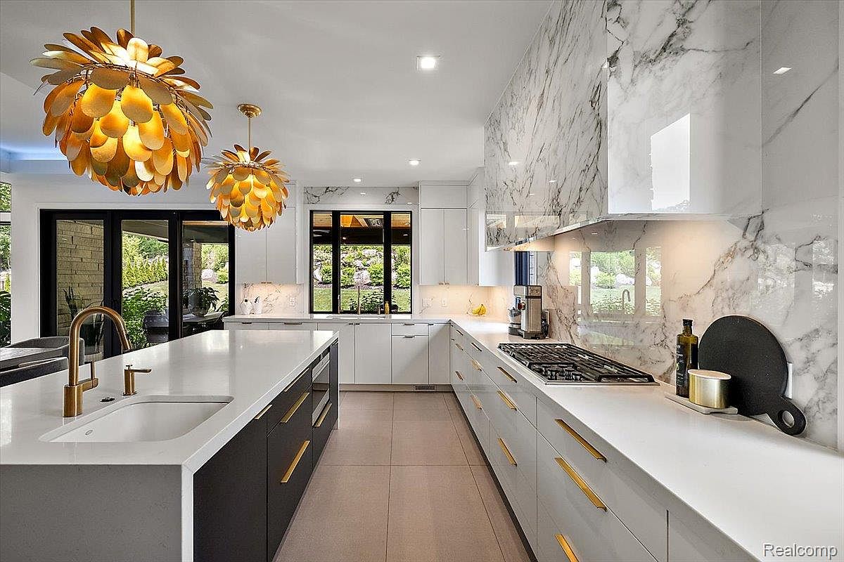 This is a bright and modern kitchen featuring a large island with a white countertop and dark gray cabinetry. The kitchen is equipped with stainless steel appliances, gold hardware, and a marble backsplash that extends to the ceiling. Large windows provide natural light and a view of the outdoors, enhancing the open and airy feel of the space.