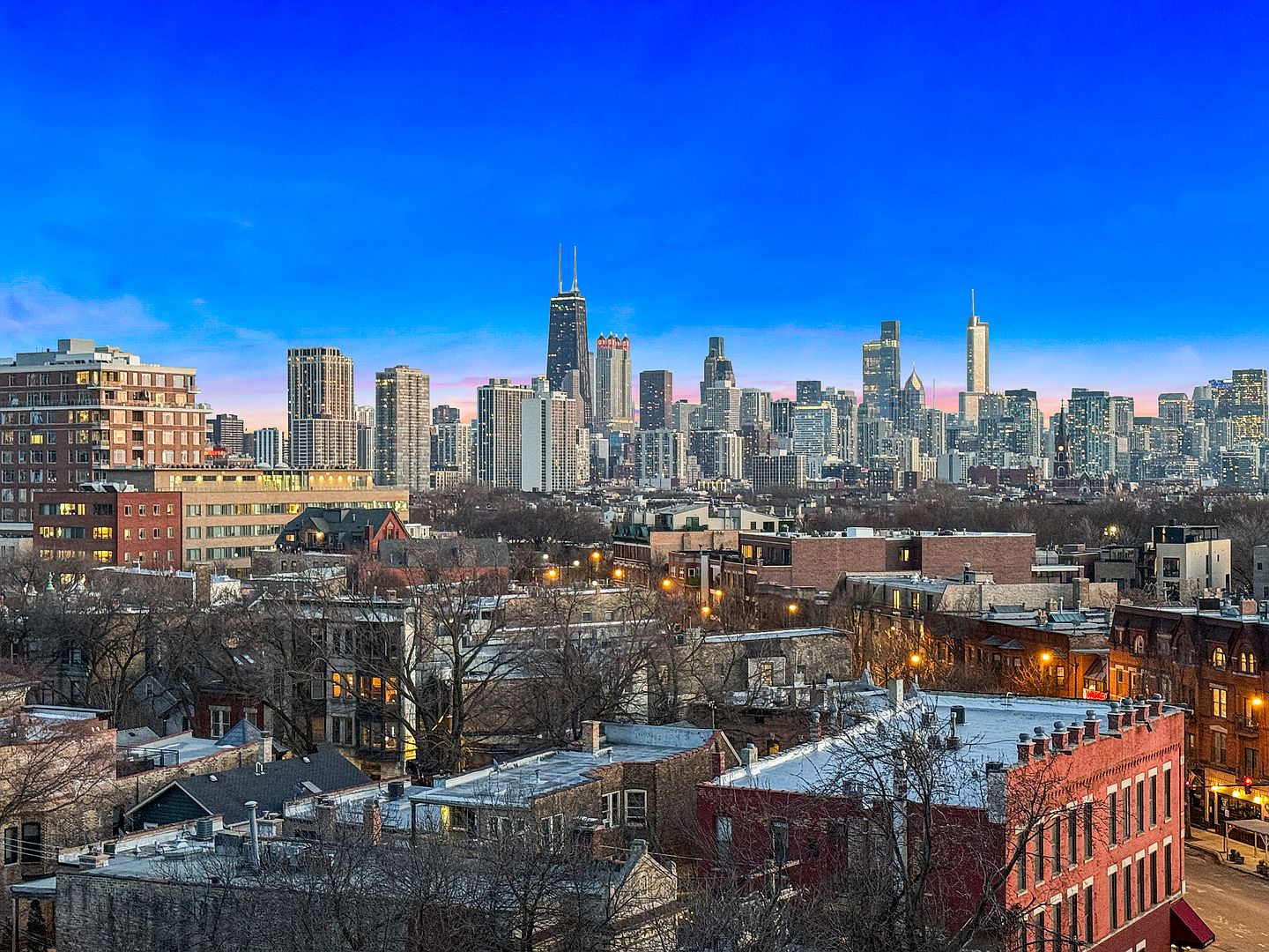 This elevated aerial view captures a sprawling cityscape at dusk, featuring a dense urban skyline dominated by iconic skyscrapers under a vibrant blue and pink gradient sky. In the foreground, the scene showcases a mix of residential brick buildings, bare-branched trees, and neighborhood rooftops, providing a sense of the property's location within a bustling metropolitan area. The perspective offers a wide, cinematic look at the city's architectural density and the transition from residential streets to the high-rise commercial core.