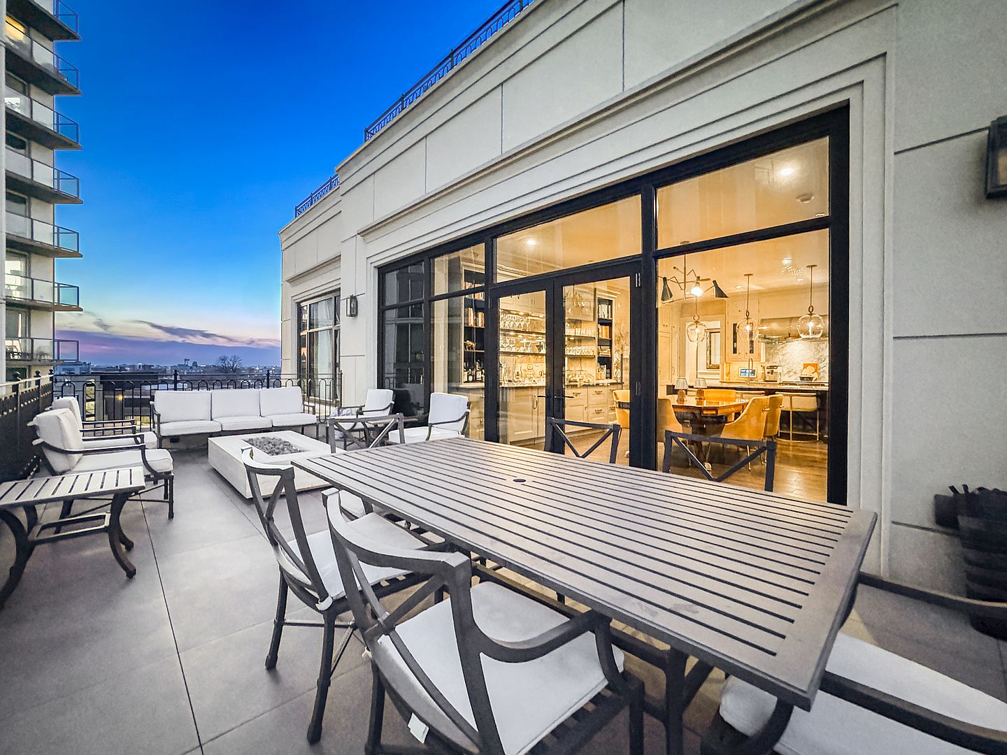 This expansive rooftop terrace features a sophisticated outdoor dining area with a long table and comfortable seating, complemented by a separate lounge area with a fire pit. Large glass doors provide a seamless transition between the outdoor space and the interior dining room, which is visible through the glazing. The setting offers a panoramic view of the city skyline at dusk, creating an elegant and inviting atmosphere for evening entertaining.