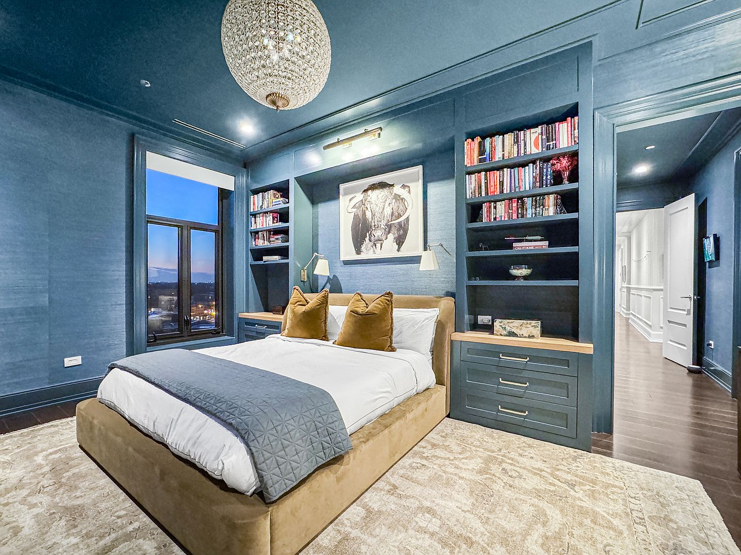 This sophisticated primary bedroom features deep blue walls and built-in shelving flanking a central bed with a velvet-textured frame. A large crystal chandelier serves as a focal point above the bed, while a framed bull artwork adds a bold artistic touch. The room is completed by a neutral patterned area rug and a window offering a view of the city skyline at dusk.