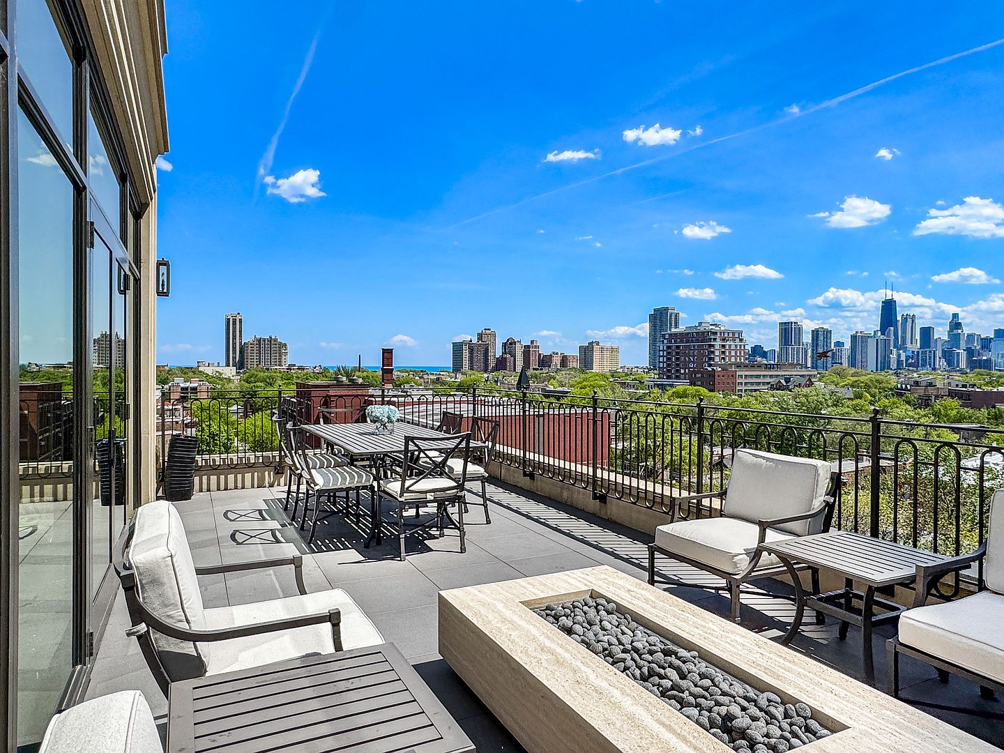 This expansive rooftop terrace offers a sophisticated outdoor living space with a panoramic view of the city skyline. The area is furnished with a dining set and comfortable lounge seating arranged around a rectangular stone fire pit filled with dark river stones. Large glass doors provide seamless access from the interior, creating an ideal setting for entertaining against a backdrop of urban architecture.