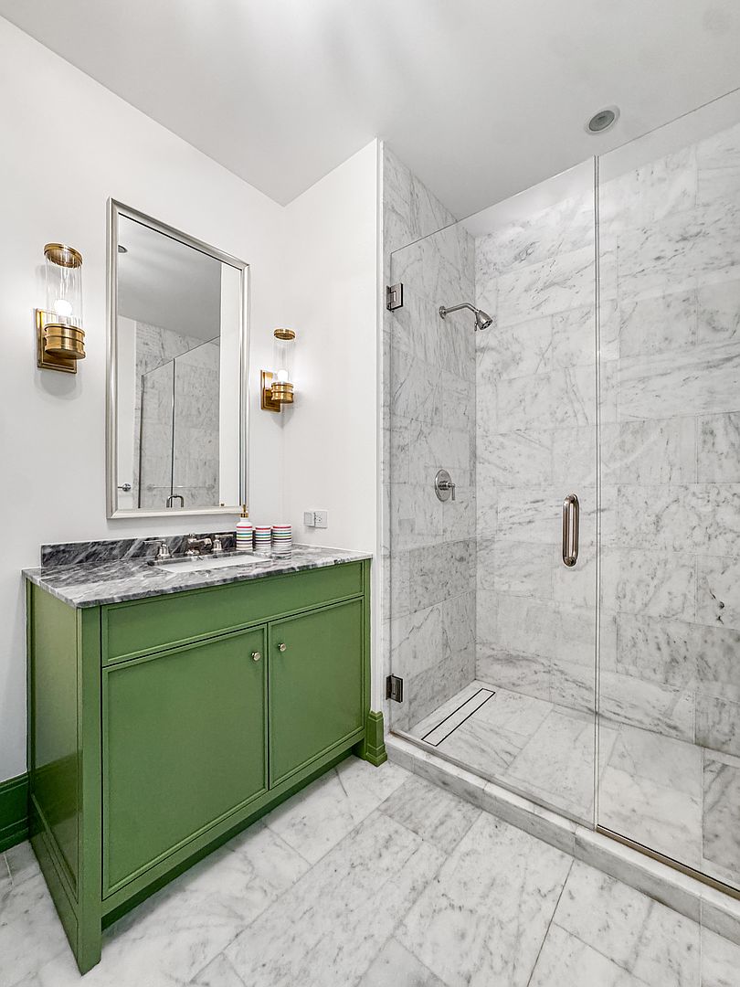 This bright bathroom features a striking green vanity topped with a grey-veined stone counter and a rectangular undermount sink. To the right, a walk-in shower is enclosed by a glass panel and finished with light-colored stone tile on both the walls and floor. The space is accented by polished metal fixtures, including wall-mounted sconces flanking a rectangular mirror, creating a clean and sophisticated aesthetic.
