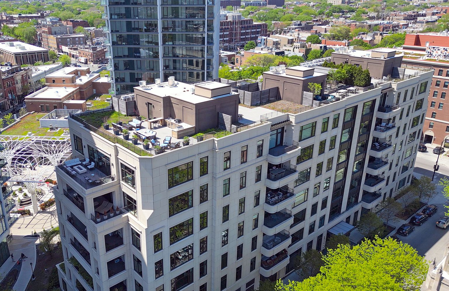 This high-angle aerial view captures a modern multi-story residential building featuring a prominent rooftop terrace with lounge seating and outdoor dining areas. The structure showcases a light-colored facade with numerous balconies and large windows, set within a dense urban neighborhood. The perspective highlights the building's integration into the surrounding cityscape, emphasizing its outdoor amenities and proximity to local streets.