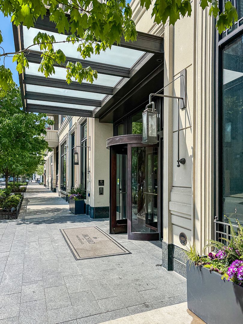 This sophisticated building entrance features a prominent revolving door framed by elegant, dark-toned metal and glass. A modern glass-and-metal canopy extends over the sidewalk, complemented by classic wall-mounted lanterns and a branded welcome mat. The exterior is finished in a light-colored, textured material, creating a polished and professional urban aesthetic.