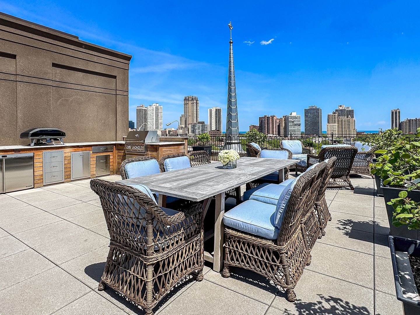 This expansive rooftop terrace offers a sophisticated outdoor dining area complete with a large wooden table, woven wicker chairs with blue cushions, and a built-in outdoor kitchen featuring stainless steel appliances. The space is set against a stunning urban skyline backdrop, providing a panoramic view of city buildings and a prominent church spire. The clean, paved surface and well-maintained greenery create an inviting atmosphere for entertaining in an elevated city setting.
