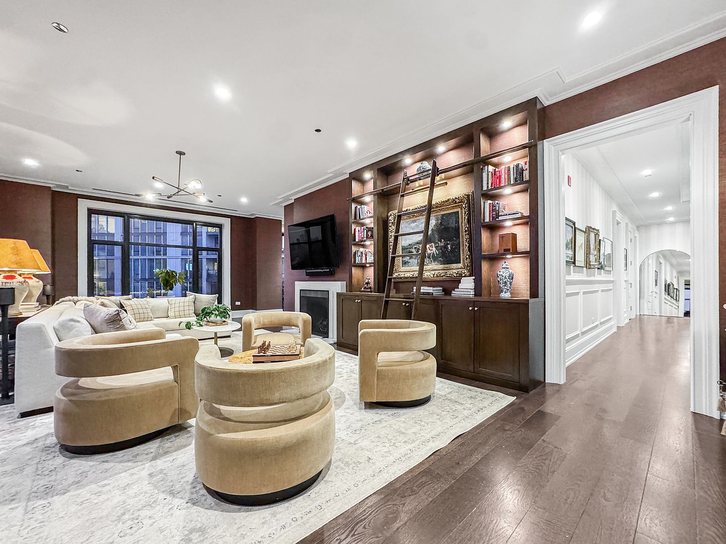 This sophisticated living room features deep, warm-toned walls and rich dark wood flooring, creating an inviting and elegant atmosphere. A large, custom-built dark wood shelving unit with an integrated ladder serves as a focal point, housing books and decorative items alongside a television mounted above a fireplace. The space is furnished with a plush white sectional sofa and several curved, neutral-toned armchairs arranged around a central area rug, while a wide opening leads into a bright, white-trimmed hallway.