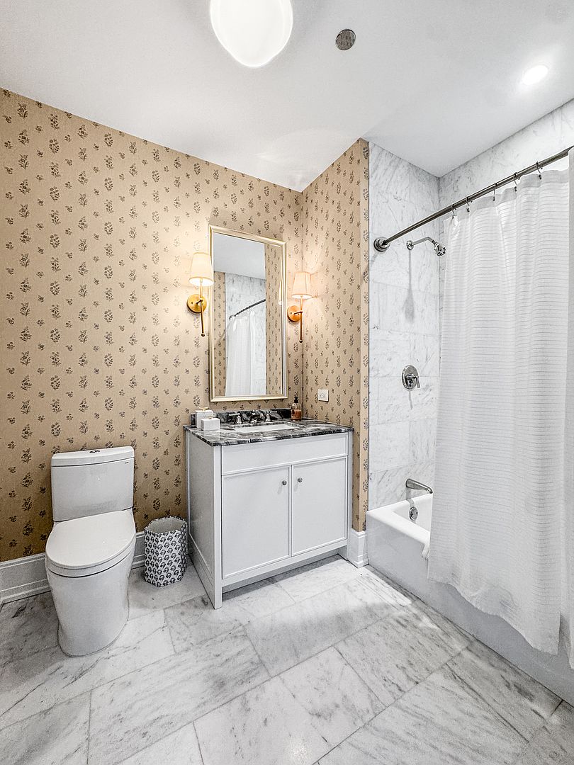 This bright, well-appointed bathroom features patterned wallpaper and light-colored stone flooring. The space includes a white vanity with a dark stone countertop, a wall-mounted mirror flanked by two sconces, and a shower-tub combination enclosed by a white curtain. The perspective is a wide-angle shot from the doorway, capturing the entire layout of the room.