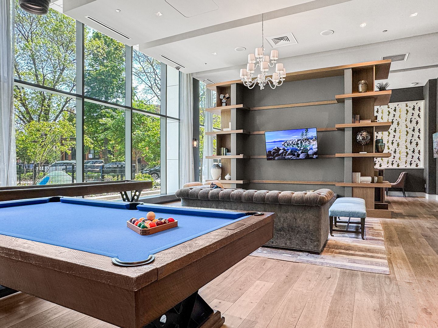 This spacious community lounge features a blue-felt pool table in the foreground and a comfortable seating area with a tufted sofa facing a wall-mounted television. The room is anchored by a large, dark-toned shelving unit and illuminated by a classic chandelier, while expansive floor-to-ceiling windows provide abundant natural light and views of the surrounding greenery. The space combines modern design with a relaxed, social atmosphere, perfect for resident gatherings.