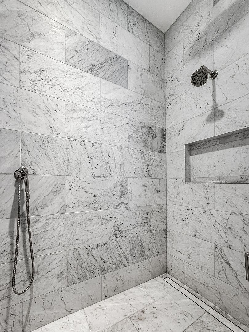 This shower enclosure features large-format stone tiles with a distinct veined pattern covering the walls and floor. The space is equipped with both a wall-mounted shower head and a handheld sprayer on a flexible hose, alongside a recessed wall niche for storage. A linear drain is integrated into the floor, contributing to the clean, minimalist aesthetic of this modern bathroom.