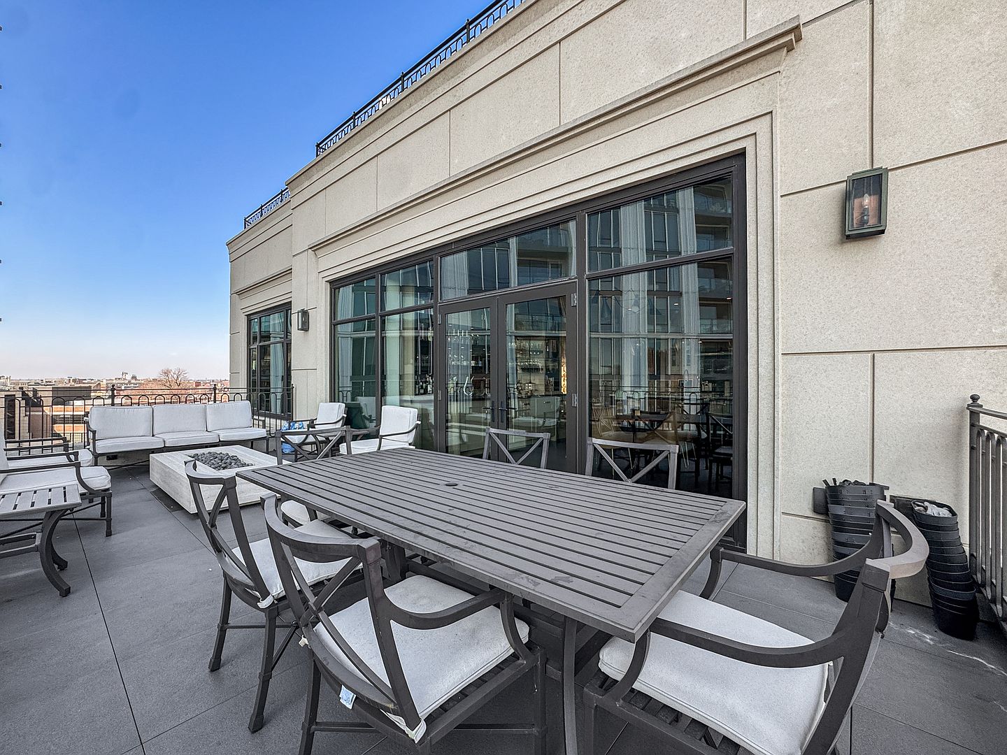 This spacious rooftop terrace features a modern outdoor dining set and a comfortable lounge area with a fire pit. Large glass doors provide a seamless transition between the indoor living space and the expansive deck, which offers a clear view of the surrounding urban landscape. The neutral stone facade and clean lines create a sophisticated, contemporary atmosphere perfect for outdoor entertaining.