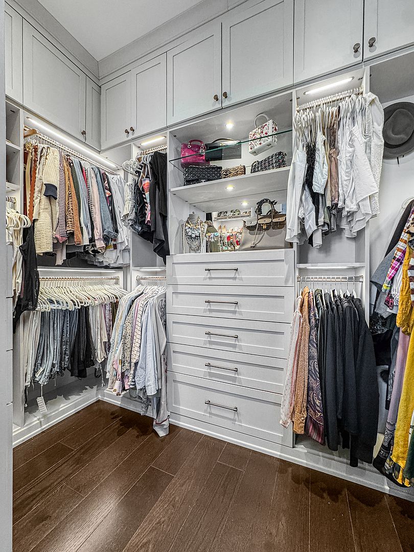 This organized walk-in closet features custom light-colored cabinetry with integrated LED lighting throughout the shelving and hanging rods. The space includes a central bank of five drawers with modern hardware, open shelving for accessories, and dark wood-toned flooring. The layout maximizes storage efficiency while maintaining a clean, high-end aesthetic.