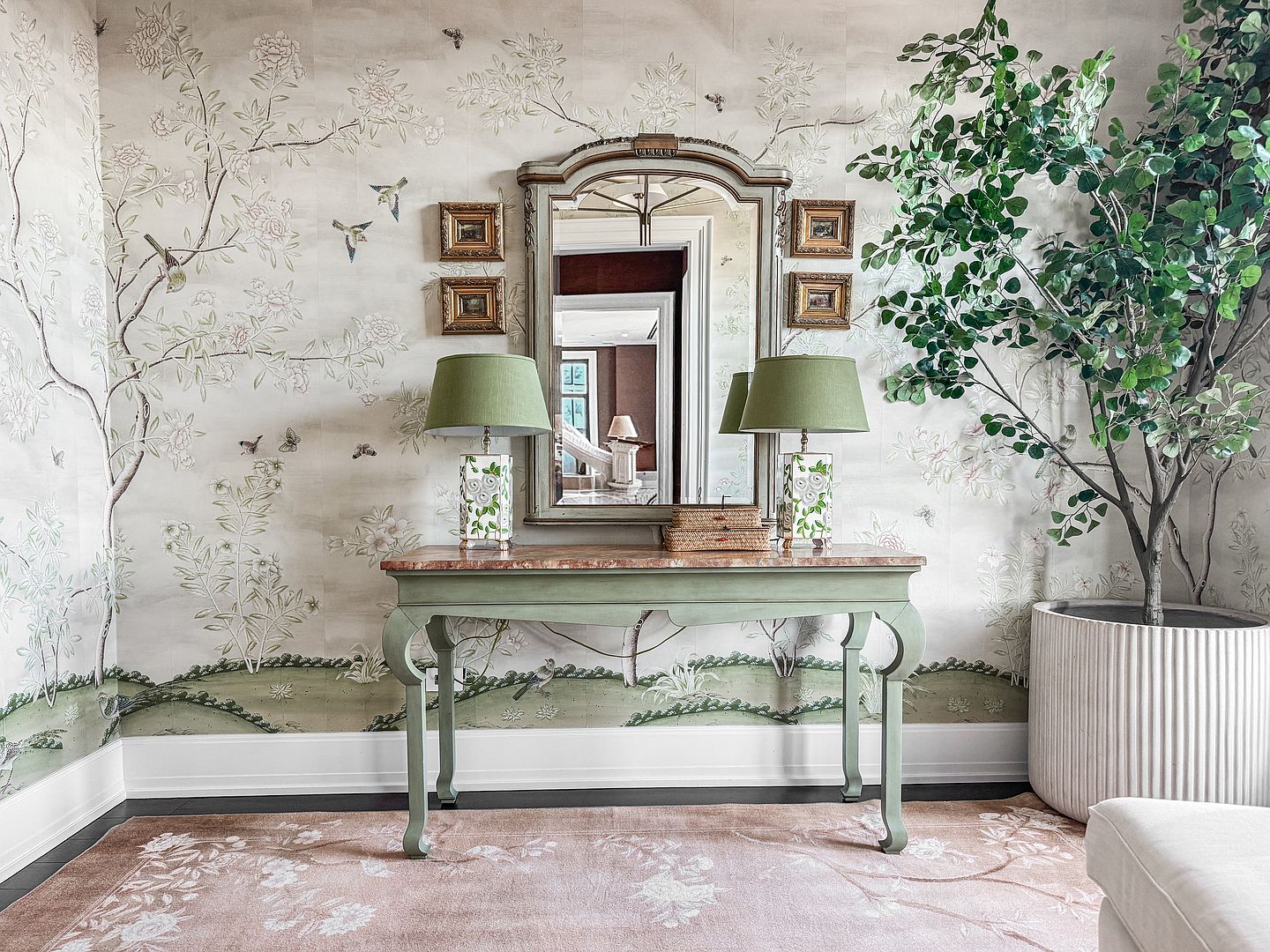 This elegant entryway features a console table with a stone top positioned against a wall adorned with intricate, nature-inspired wallpaper. A central mirror is flanked by two pairs of small framed artworks, while matching green lamps provide soft lighting on either side of the table. A large potted tree adds a touch of greenery to the corner, and a patterned rug anchors the space, creating a sophisticated and welcoming atmosphere.