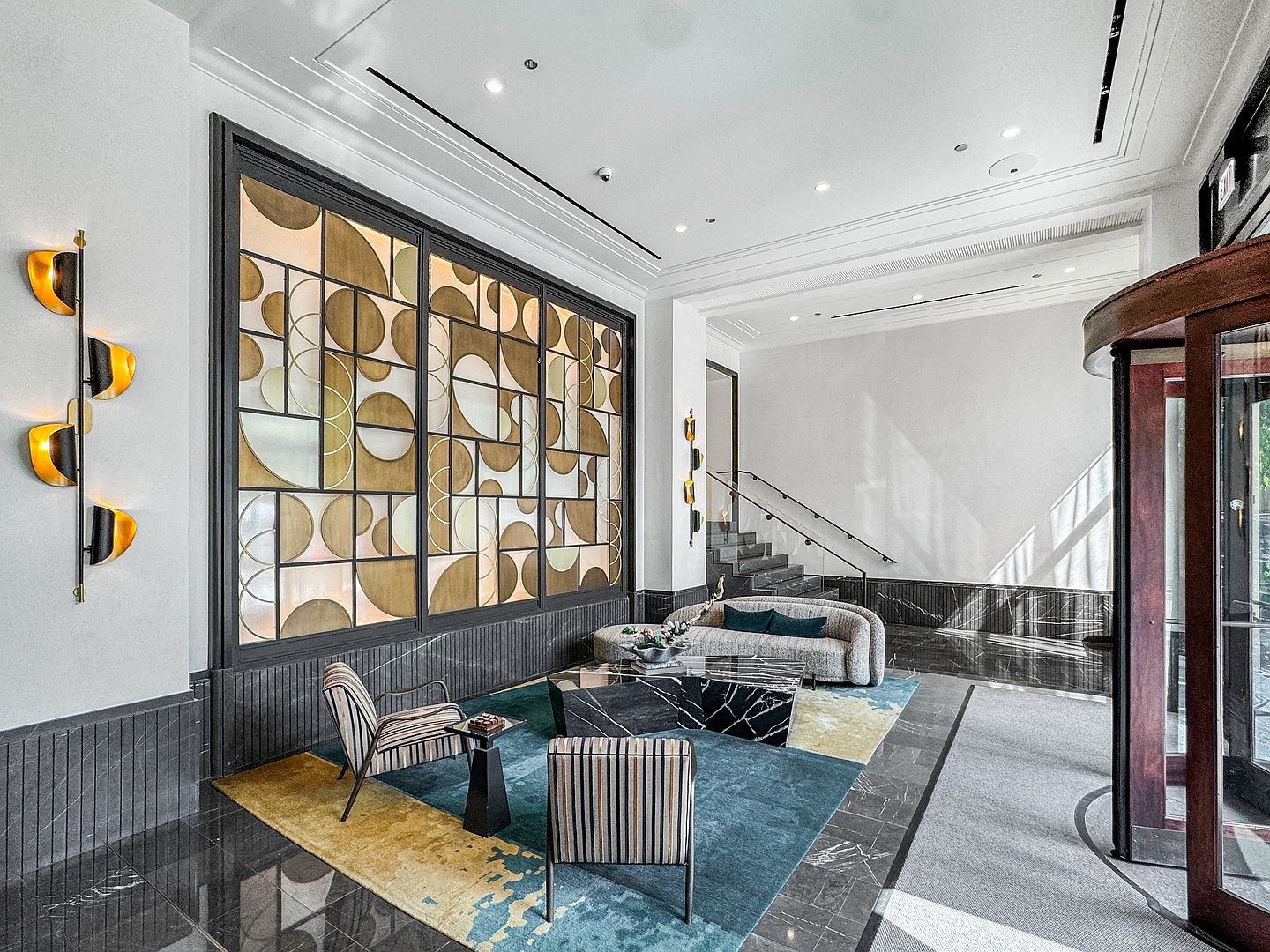 This sophisticated lobby or building entrance features a striking geometric art installation on the wall, complemented by a modern seating area with a curved sofa and patterned armchairs. The space is anchored by a vibrant, multi-colored area rug and a dark stone coffee table, set against a backdrop of dark stone flooring and wall paneling. A revolving door on the right leads to the exterior, while a staircase with a sleek metal railing adds architectural depth to the refined, upscale interior.