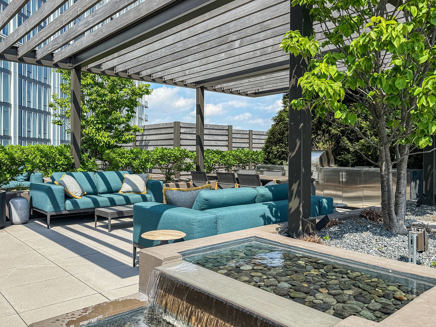 This sophisticated rooftop terrace features a modern pergola providing shade over a comfortable seating area with teal-colored outdoor sofas. A tranquil water feature with smooth river stones serves as a focal point in the foreground, while a stainless steel outdoor kitchen and dining area are positioned nearby. The space is surrounded by lush greenery and contemporary fencing, creating a serene and upscale urban retreat.