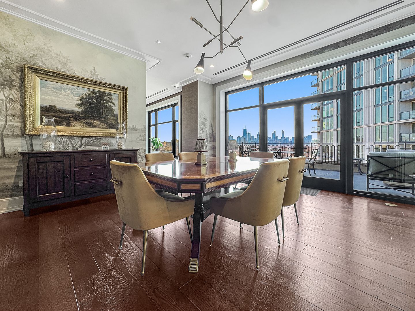 This elegant dining room features a polished wood table surrounded by six upholstered chairs, set against a backdrop of mural-style wallpaper and a large landscape painting. The space is illuminated by a modern, multi-armed chandelier and benefits from abundant natural light streaming through large windows that offer a clear view of the city skyline. The room is anchored by a dark wood sideboard and rich, warm-toned wood flooring, creating a sophisticated and inviting atmosphere.