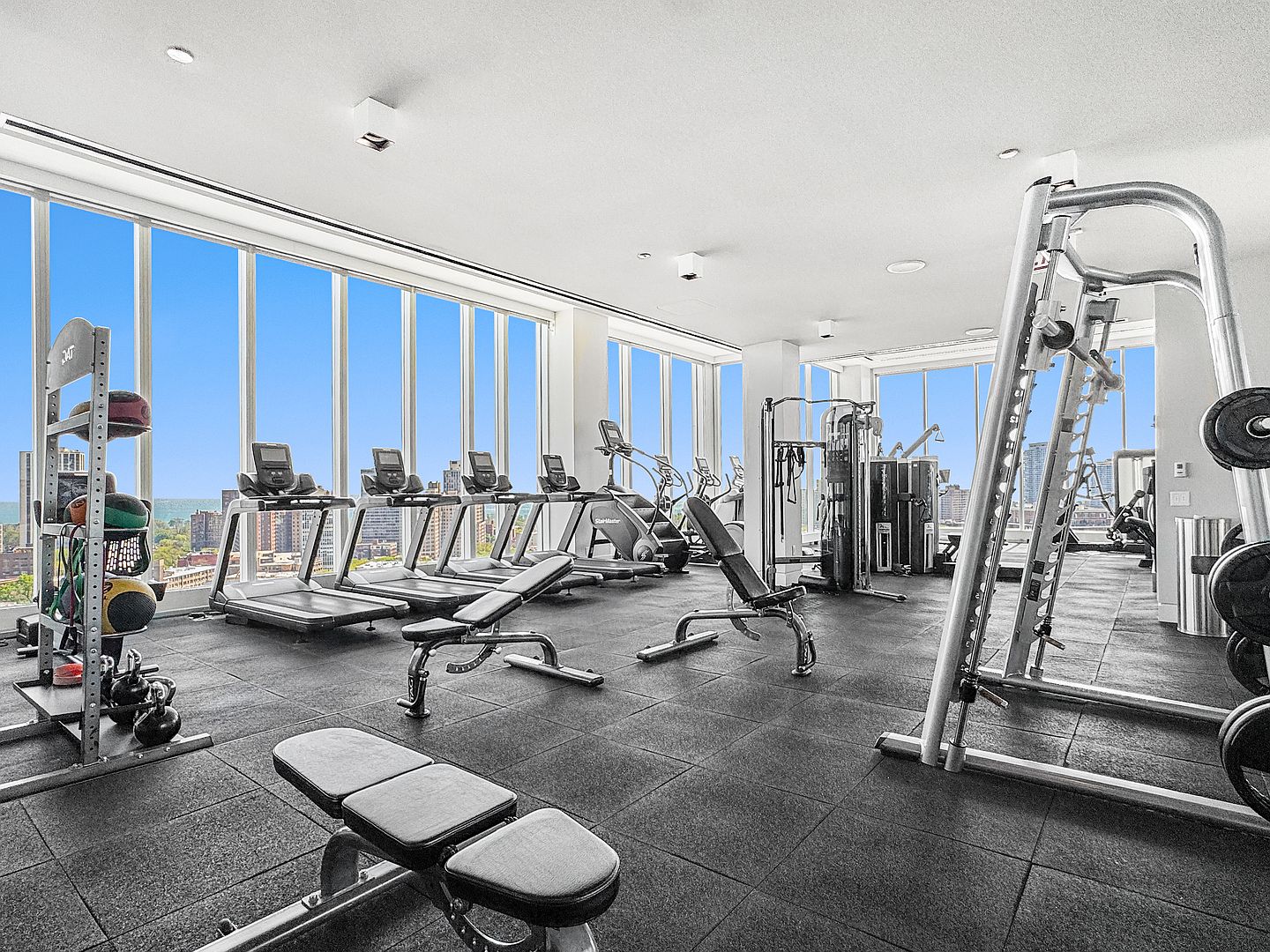 This expansive, modern fitness center features a wide array of professional-grade equipment, including treadmills, a Smith machine, and adjustable weight benches. The space is defined by its extensive floor-to-ceiling windows that offer panoramic city views and flood the room with natural light. The dark, durable rubber flooring provides a sleek, functional foundation for the bright and airy workout environment.