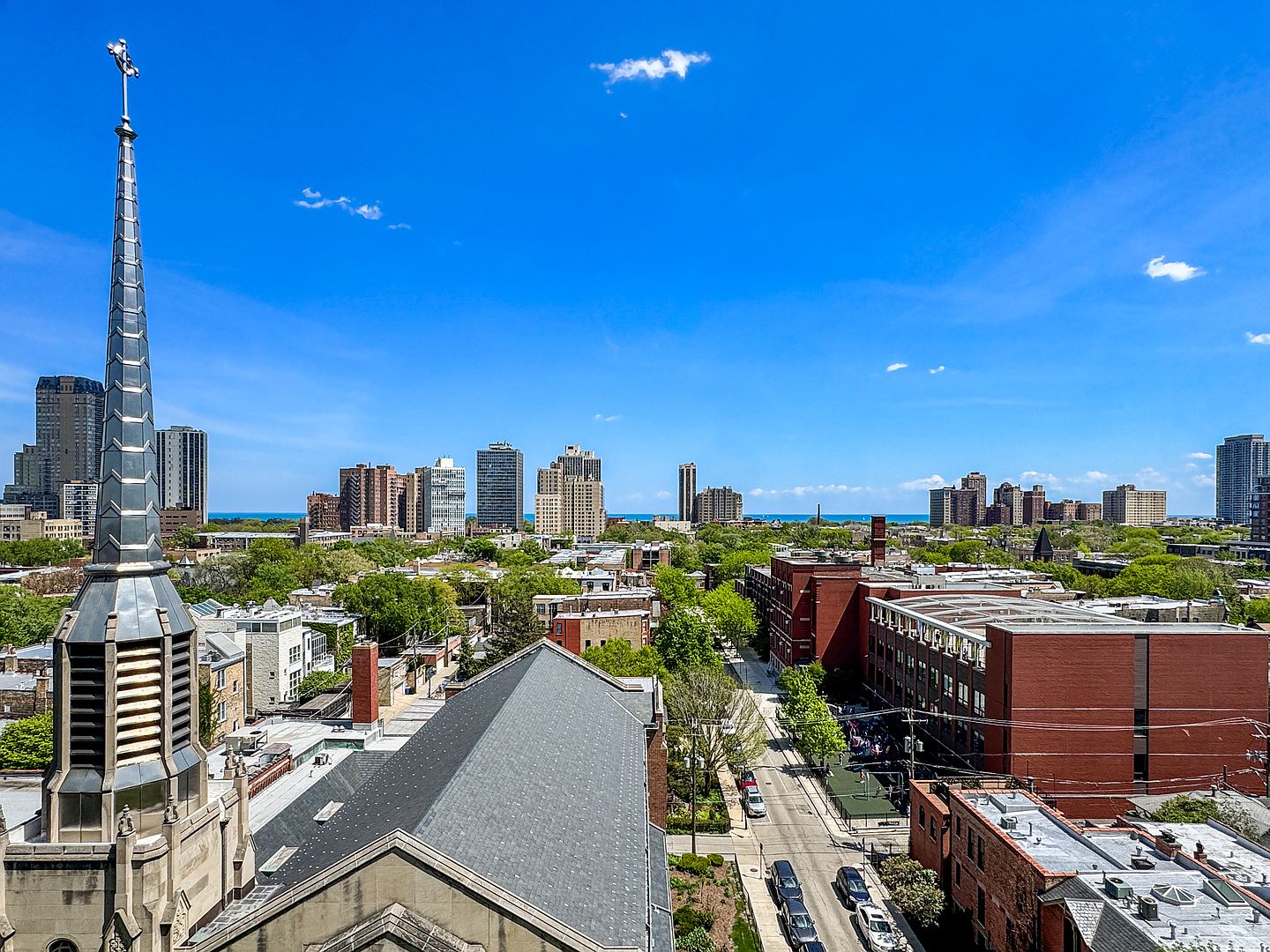 This high-angle aerial view captures a cityscape featuring a prominent church steeple in the foreground and a dense urban neighborhood leading toward a distant skyline and the horizon. The scene highlights the architectural contrast between the historic church roof and the surrounding brick buildings, tree-lined streets, and modern high-rise towers. The bright blue sky and clear visibility emphasize the property's prime location within a vibrant metropolitan area.