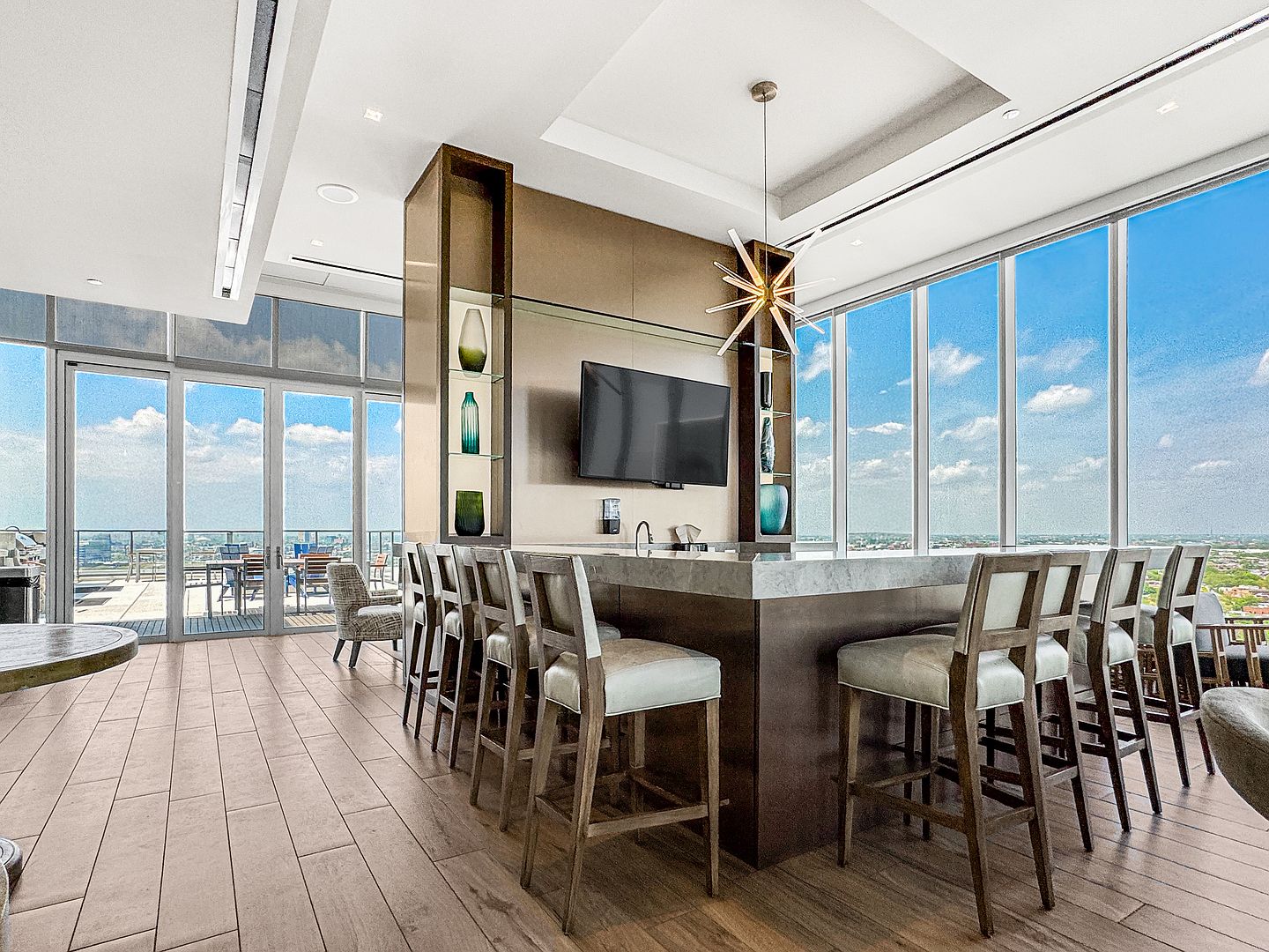 This sophisticated lounge area features a large stone-topped bar with multiple stools, perfect for social gatherings. The space is defined by a central pillar with built-in shelving and a mounted television, complemented by a striking starburst-style chandelier. Large windows provide expansive views of the city skyline and access to an adjacent outdoor terrace, creating an airy and upscale atmosphere.