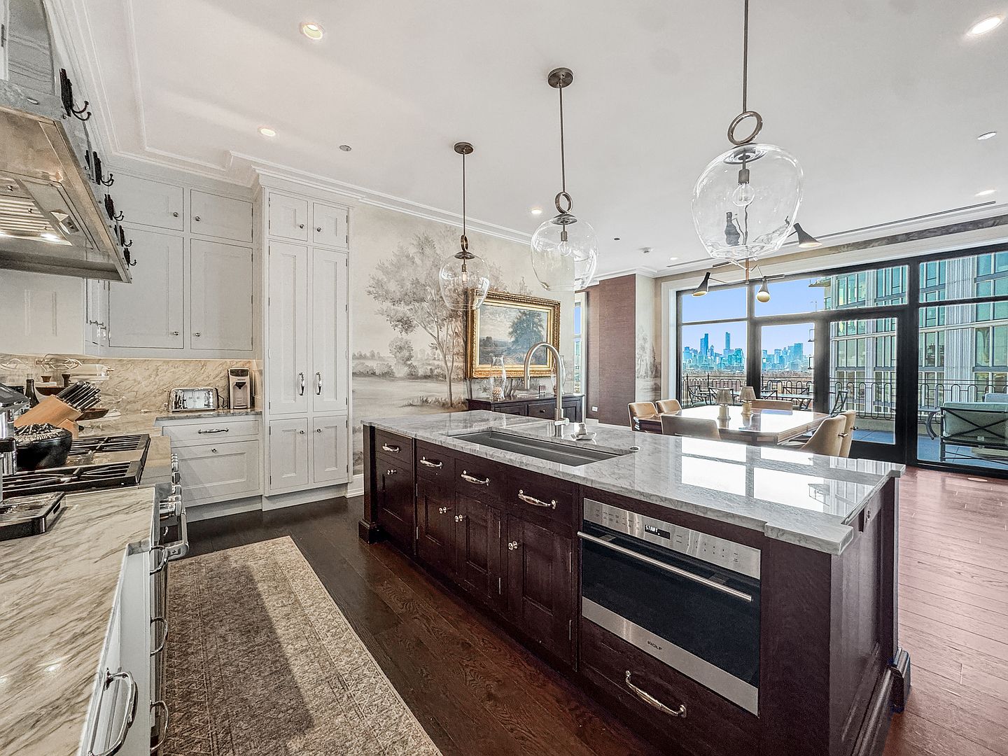 This sophisticated kitchen features a large, dark-toned central island with a stone countertop and integrated appliances, contrasting against light-colored perimeter cabinetry. A mural-style wallpaper adds an artistic touch to the wall, while large glass doors provide a seamless transition to an outdoor balcony with city views. The space is illuminated by elegant glass pendant lights, creating a refined and open atmosphere suitable for both cooking and entertaining.