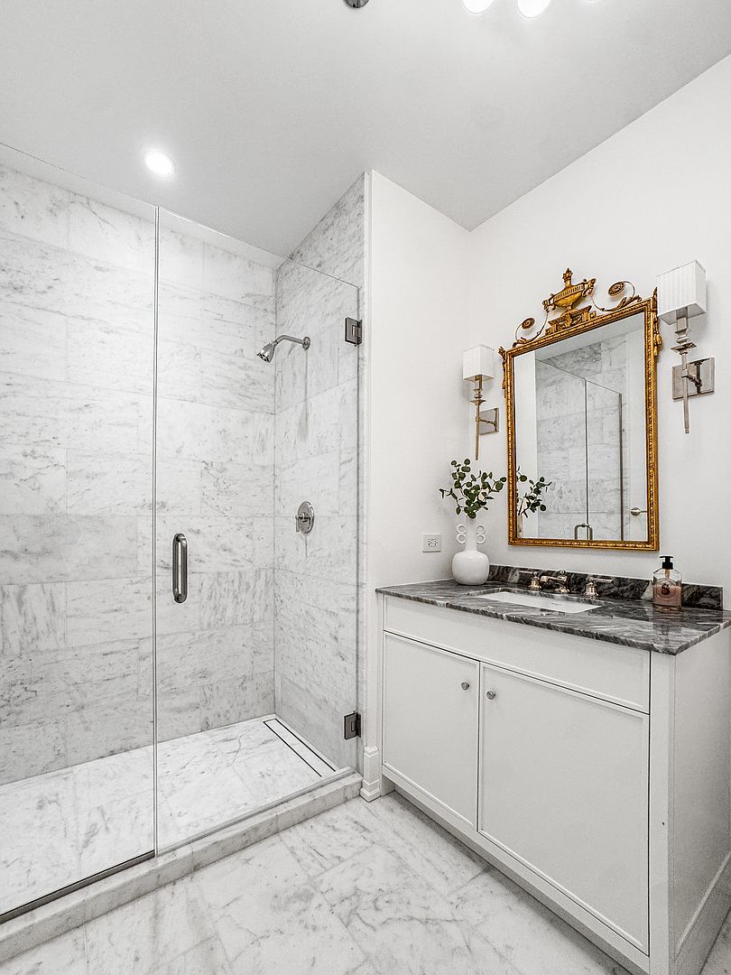 This elegant bathroom features a walk-in shower enclosed by a glass panel, showcasing light-toned stone wall and floor tiles. A white vanity with a dark stone countertop and gold-toned fixtures provides a sophisticated contrast, complemented by an ornate gold-framed mirror. The space is well-lit with wall-mounted sconces, creating a bright and polished atmosphere.