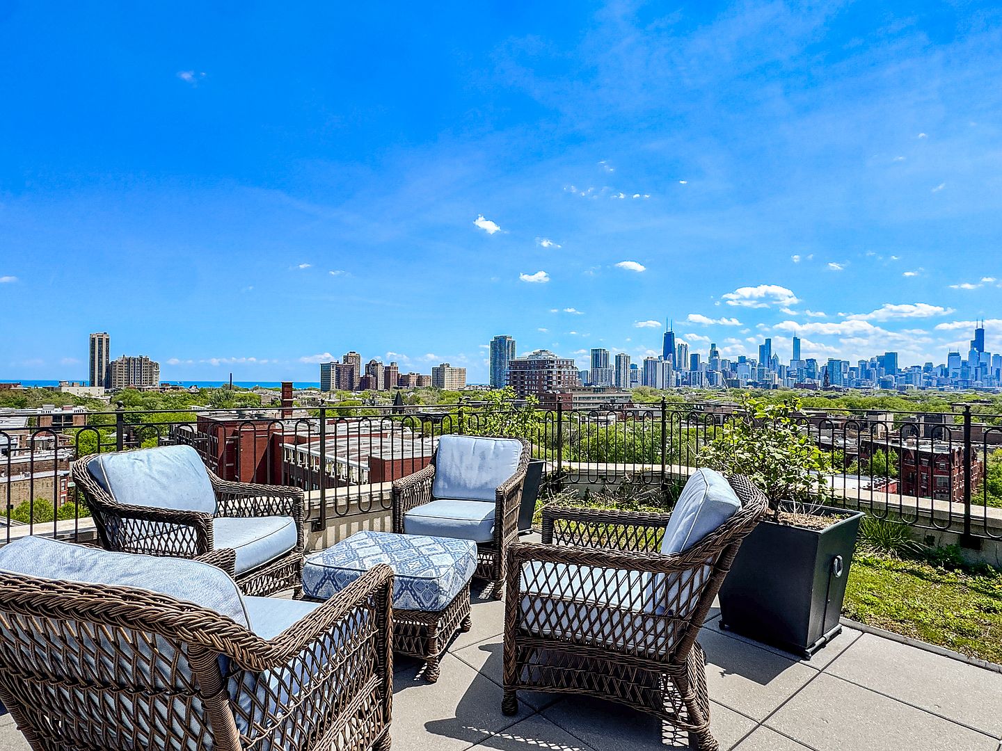 This rooftop terrace offers a stunning panoramic view of the city skyline under a bright blue sky. The space is furnished with comfortable wicker-style armchairs and a matching ottoman featuring light blue patterned cushions, creating an inviting outdoor lounge area. A black metal railing borders the deck, which also includes a large potted plant, making it an ideal spot for relaxation or entertaining.