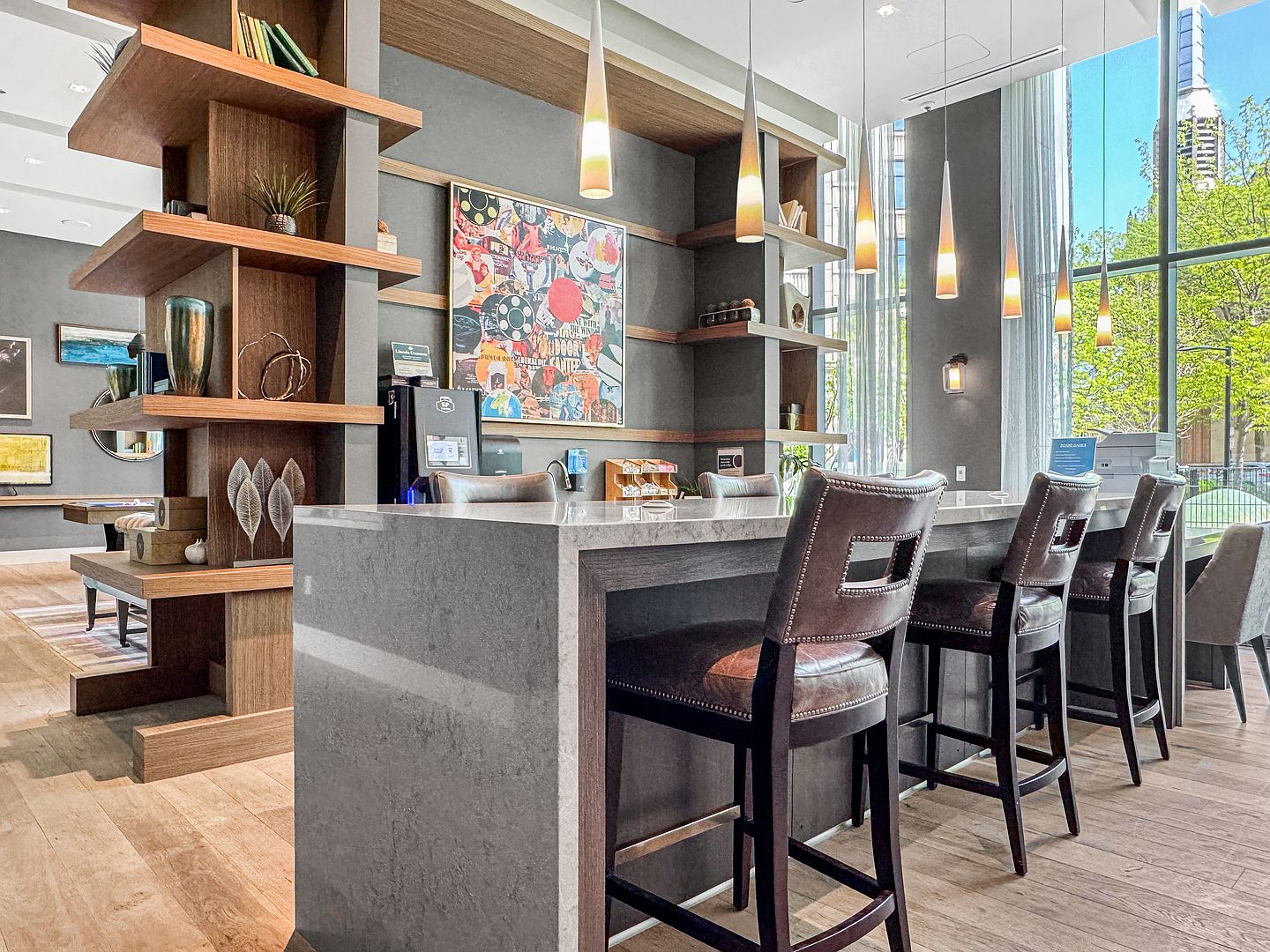 This sophisticated community lounge area features a long, stone-topped bar with leather-upholstered stools, perfect for social gatherings. The space is accented by a large, colorful abstract art piece, open shelving with decorative items, and a series of sleek, modern pendant lights. Large windows provide ample natural light and a view of the surrounding greenery, creating an inviting and upscale atmosphere.