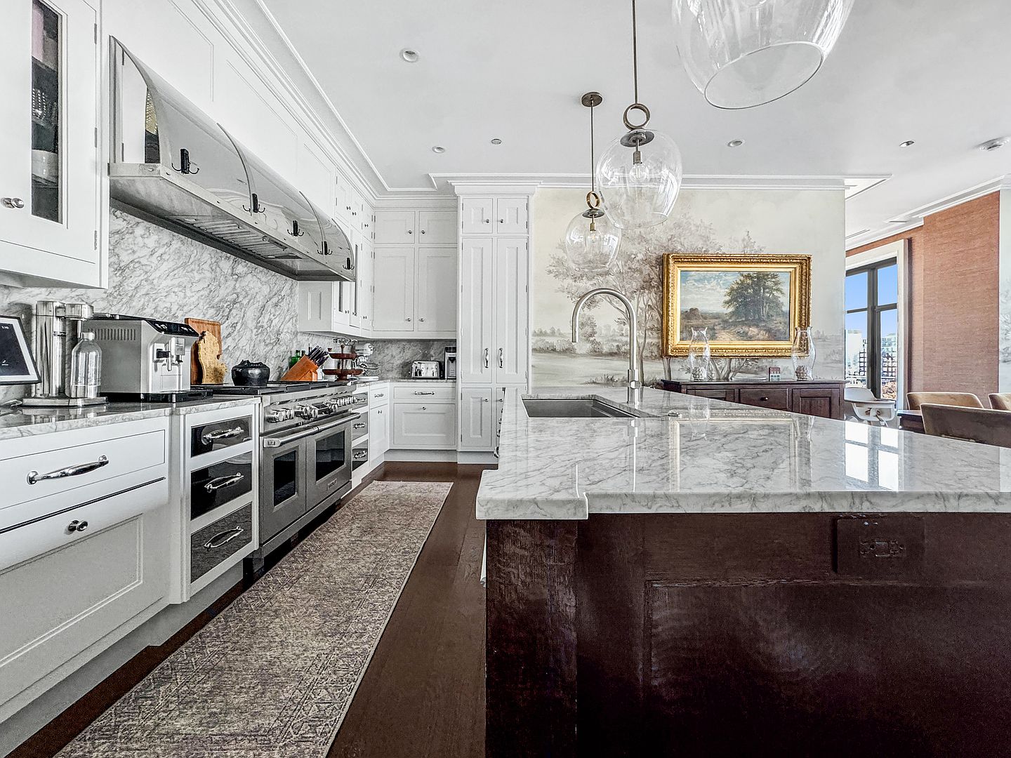 This elegant kitchen features a large central island with a stone countertop and a dark base, complemented by white cabinetry and a professional-grade stainless steel range. A mural-style wall treatment behind the island adds a sophisticated artistic touch, while glass pendant lights hang above the workspace. The perspective captures the depth of the room, showcasing the contrast between the light-toned stone surfaces and the rich, dark wood flooring.