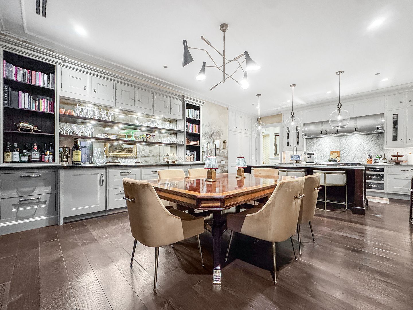 This elegant dining area features a polished wood table surrounded by upholstered chairs, set against a backdrop of custom cabinetry and a mirrored bar display. The space is illuminated by a modern multi-arm chandelier and flows seamlessly into an open-concept kitchen with a large island and stone-topped counters. Dark wood flooring anchors the room, creating a sophisticated and cohesive atmosphere suitable for both formal dining and casual entertaining.
