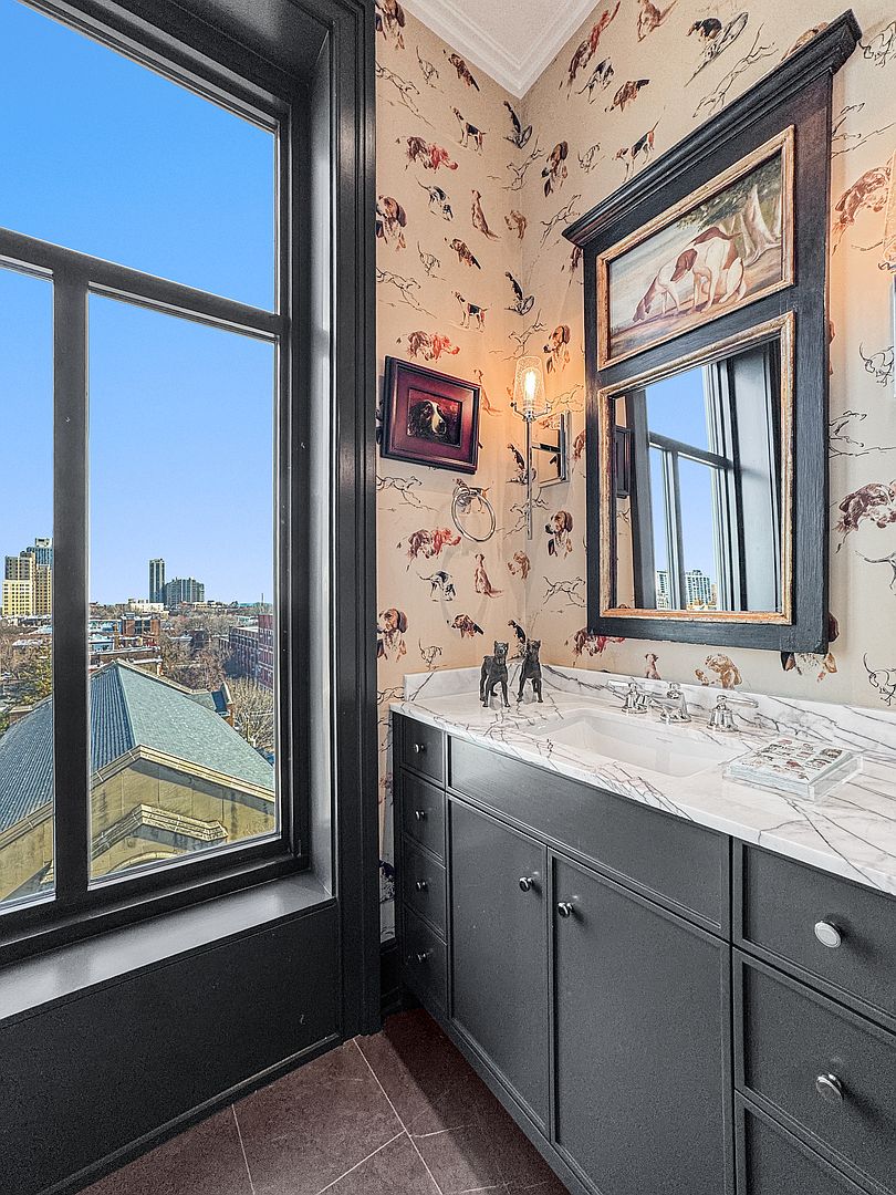 This sophisticated powder room features whimsical dog-themed wallpaper and a dark-toned vanity topped with a light-colored stone counter. A large window provides an expansive urban view, while a framed mirror and artwork add a curated, traditional touch. The space is illuminated by a wall-mounted light fixture, creating a polished and character-filled atmosphere.