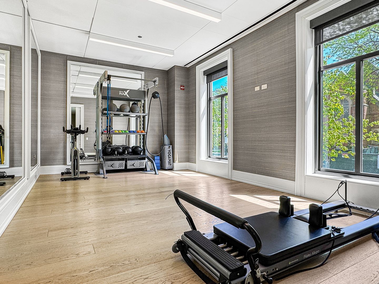 This bright and well-equipped home gym features light wood flooring and textured gray walls, creating a clean and professional atmosphere. The space is furnished with a Pilates reformer in the foreground, a stationary bike, and a comprehensive storage rack filled with various fitness equipment. Large windows allow ample natural light to fill the room, while a full-length mirror on the wall enhances the sense of space.