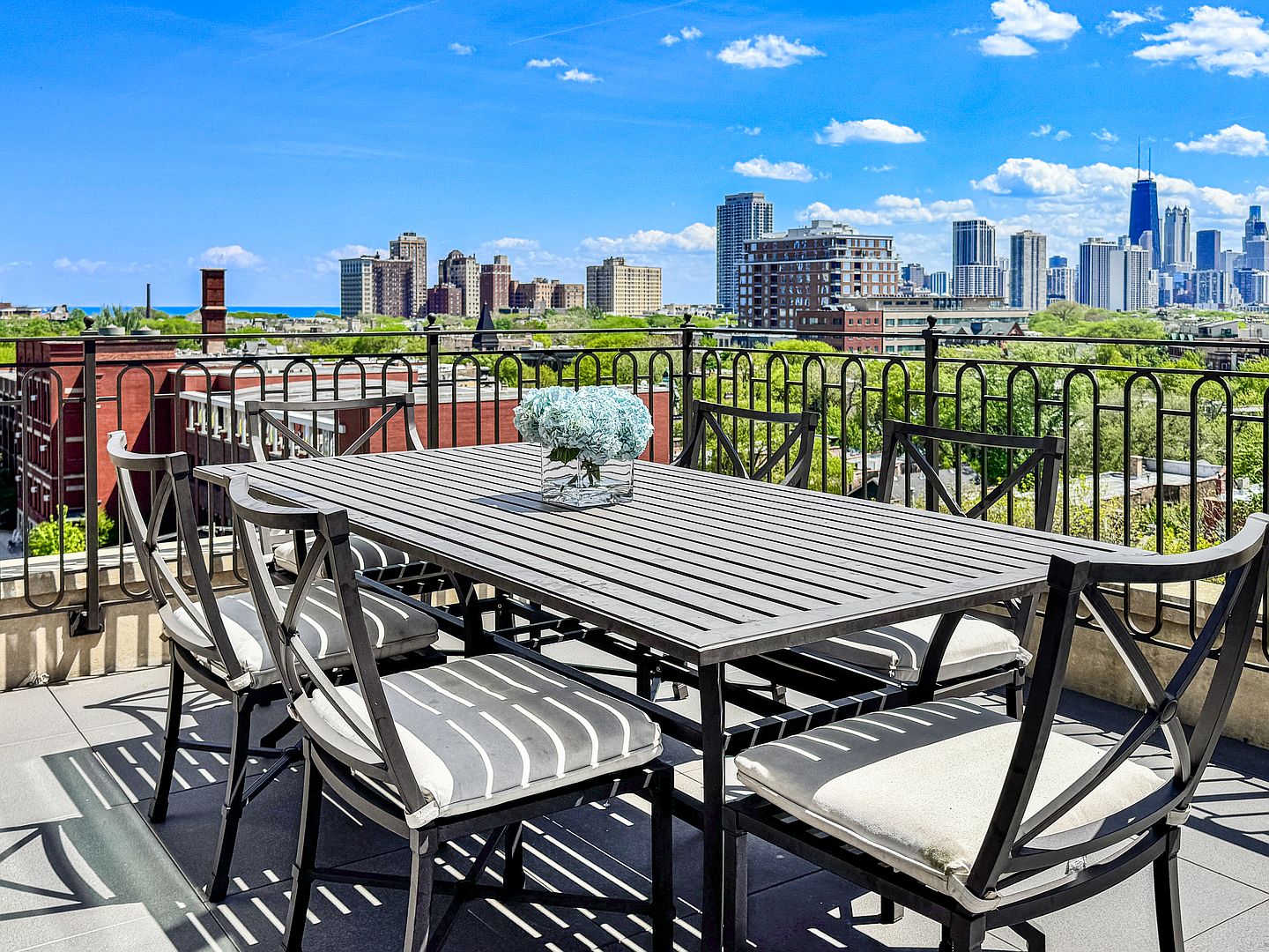 This rooftop terrace features a dark metal dining set with striped cushions, centered around a vase of blue hydrangeas. The space is enclosed by a decorative black metal railing, offering an expansive, unobstructed panoramic view of the city skyline under a bright blue sky. The perspective is eye-level, capturing the inviting outdoor dining area against the backdrop of urban architecture.