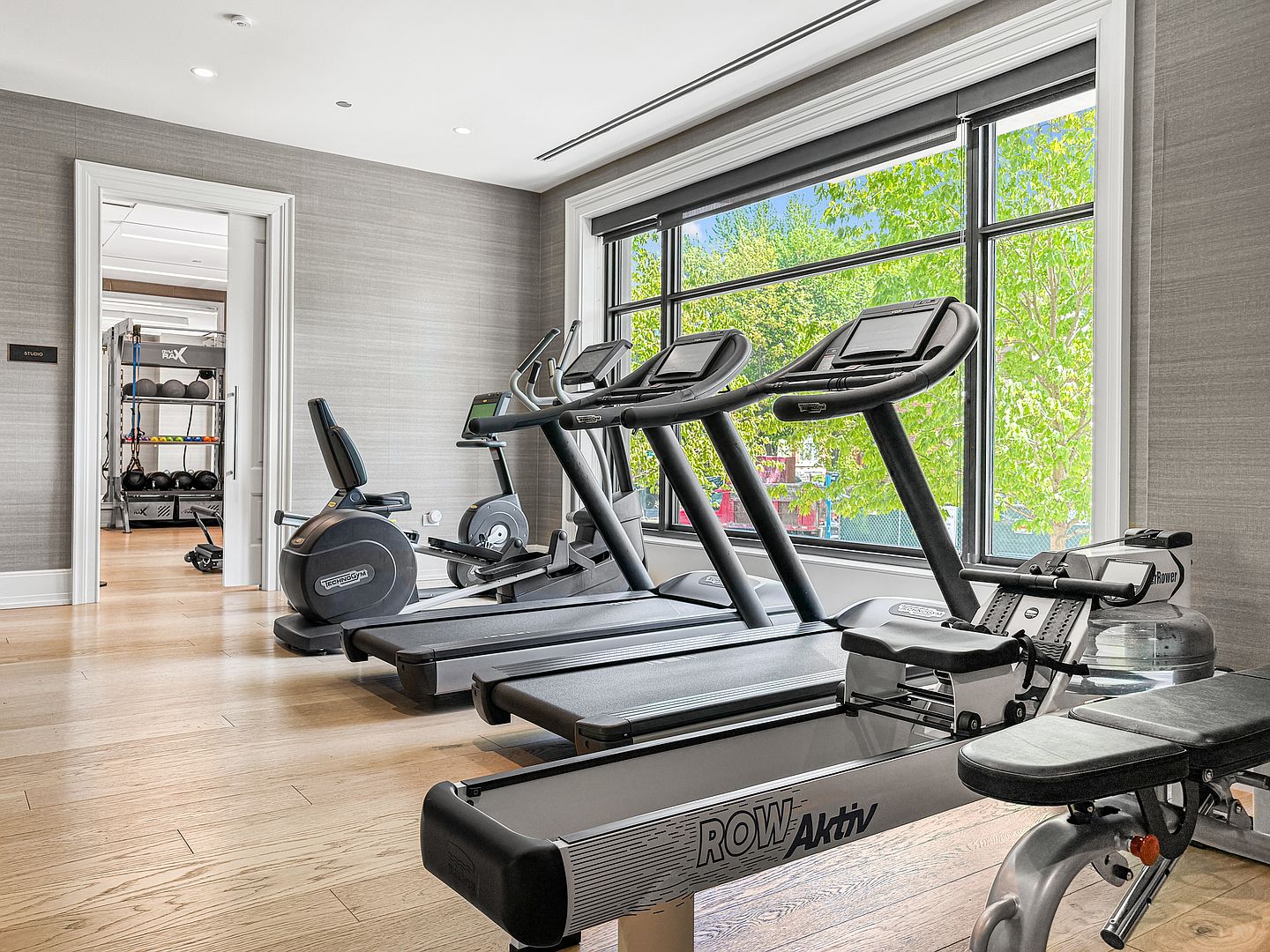 This bright and modern home gym features light wood flooring and neutral textured walls, creating a clean and professional workout environment. The space is equipped with a row of Technogym treadmills, a stationary bike, and a rowing machine positioned near large windows that provide ample natural light and a view of the outdoors. An open doorway leads to an adjacent studio area, which is stocked with various fitness equipment and storage racks.