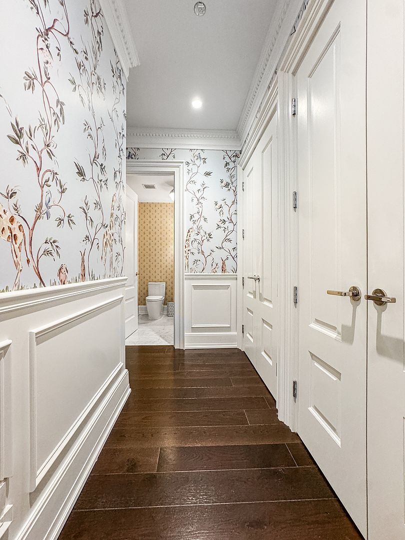 This elegant hallway features dark wood flooring and sophisticated wainscoting topped with whimsical, nature-inspired wallpaper. The perspective looks down the corridor toward a bright, neutral-toned bathroom, while white paneled doors line the right side of the space. The combination of classic architectural molding and playful wall coverings creates a refined yet inviting atmosphere.