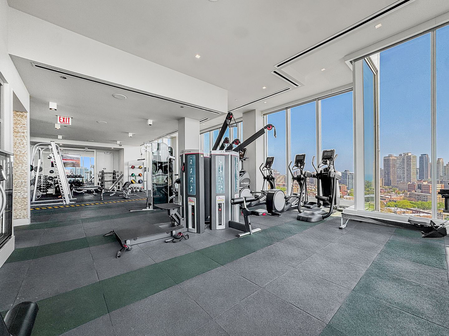 This modern, high-rise home gym features a variety of professional-grade fitness equipment, including weight machines, ellipticals, and a rack of dumbbells. The space is defined by its expansive floor-to-ceiling windows that offer a panoramic view of the city skyline. The room is finished with dark, durable rubber flooring, creating a clean and functional environment for exercise.