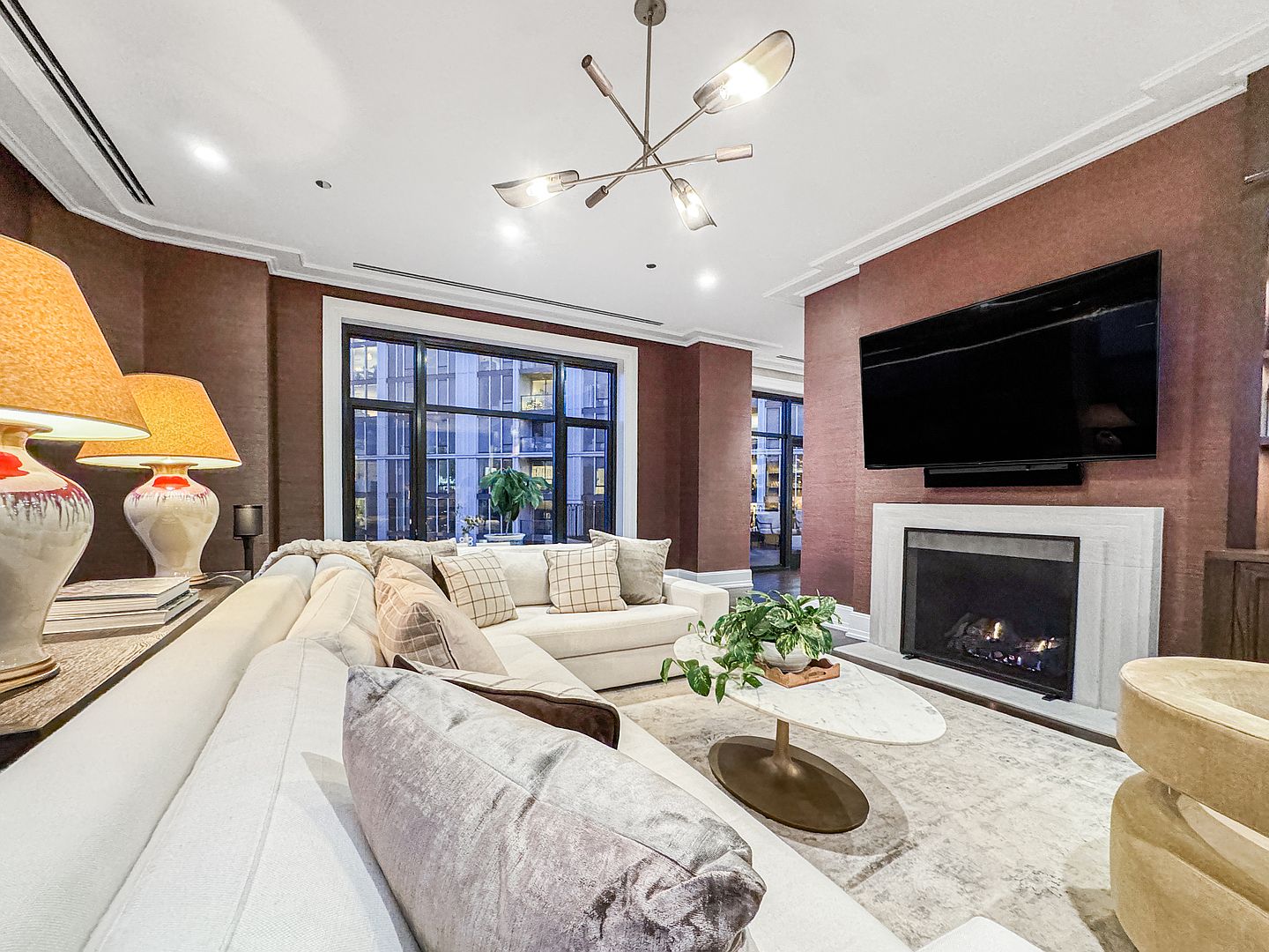 This sophisticated living room features deep, textured brown walls that create a warm and intimate atmosphere. A large, light-colored sectional sofa anchors the space, complemented by a marble-topped coffee table and a modern, multi-armed chandelier. A fireplace with a stone surround serves as a focal point, positioned beneath a wall-mounted television, while large windows provide a view of the surrounding urban architecture.