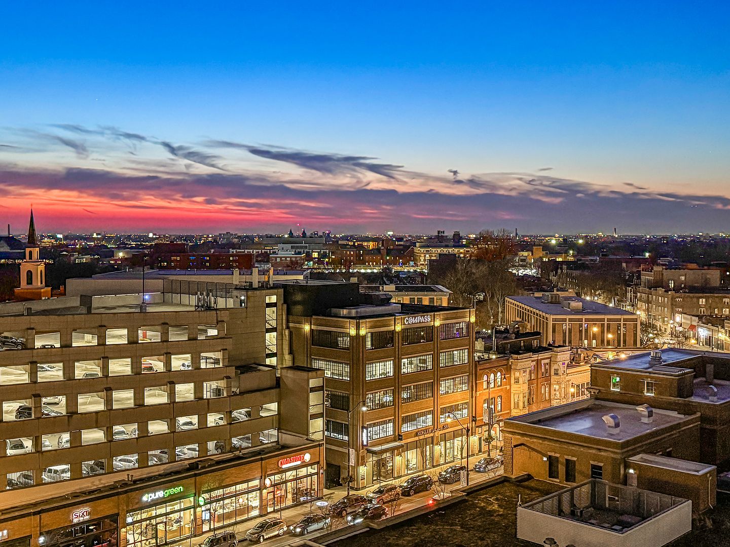 This high-angle aerial view captures a vibrant urban streetscape at dusk, featuring a multi-level parking structure and a prominent brick building marked with 'COMPASS' signage. The scene is set against a dramatic sunset sky with deep orange and blue hues, highlighting the surrounding city architecture and street-level storefronts like 'pure green' and 'Cold Stone'. The perspective provides a comprehensive look at the neighborhood's density and evening atmosphere, emphasizing the property's central location.