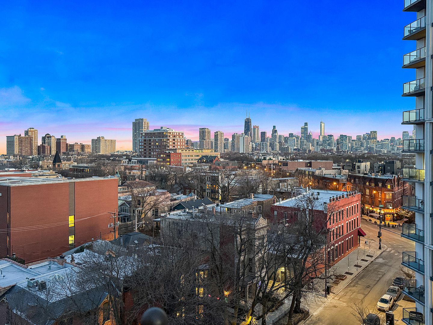 This elevated perspective captures a sweeping cityscape view of Chicago, showcasing a dense urban neighborhood in the foreground transitioning into a prominent skyline. The scene is framed on the right by the balconies of a high-rise building, while the foreground features a mix of brick residential structures and tree-lined streets. The vibrant sunset sky provides a dramatic backdrop, highlighting the architectural diversity and urban density of the area.