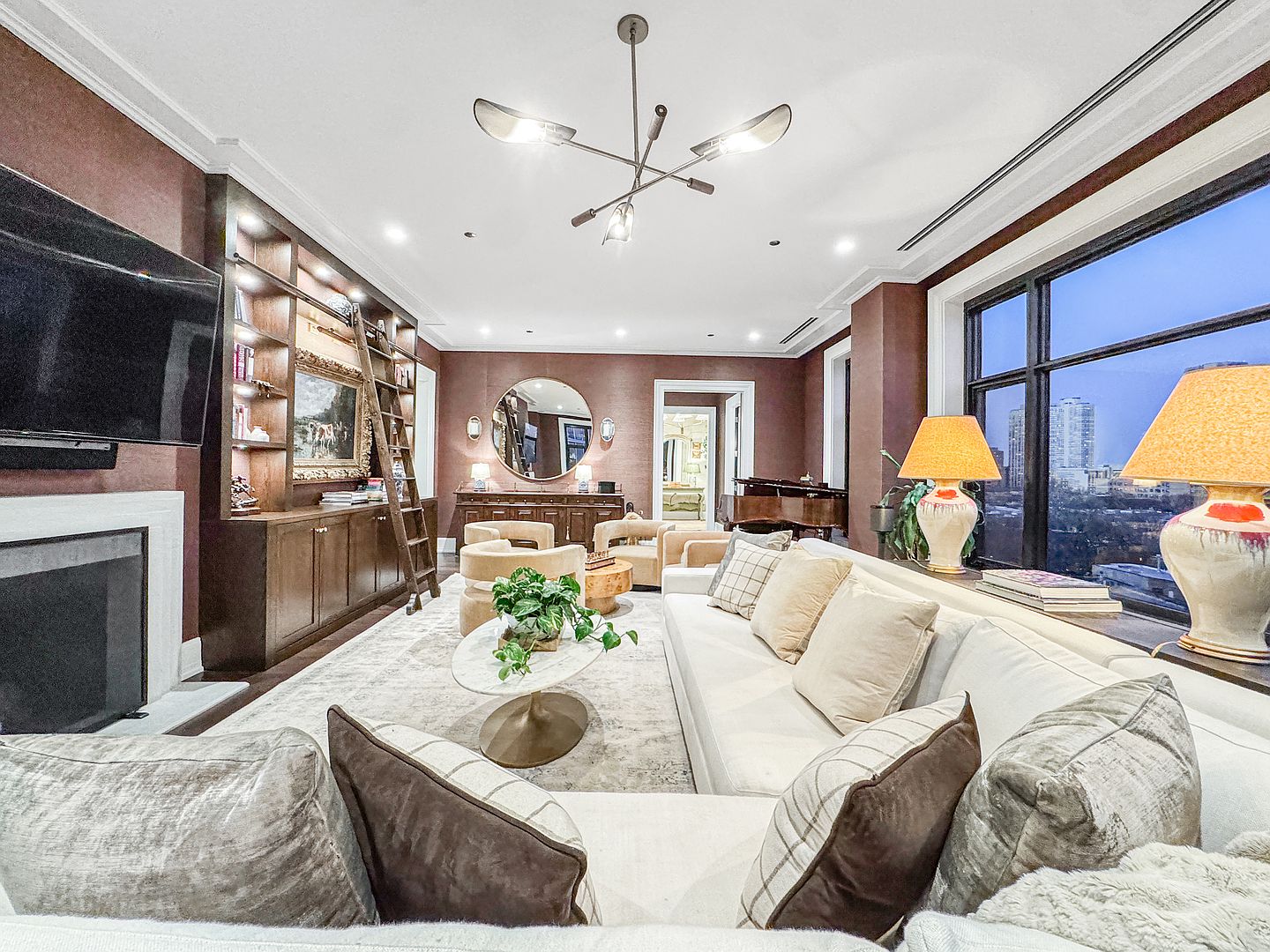 This sophisticated living room features deep, warm-toned walls and a large, light-colored sectional sofa arranged around a central marble-topped coffee table. A prominent built-in shelving unit with an integrated library ladder and a fireplace surround anchors one side of the room, while large windows offer an expansive city view on the other. The space is illuminated by a modern, multi-armed chandelier and elegant table lamps, creating a luxurious and inviting atmosphere.