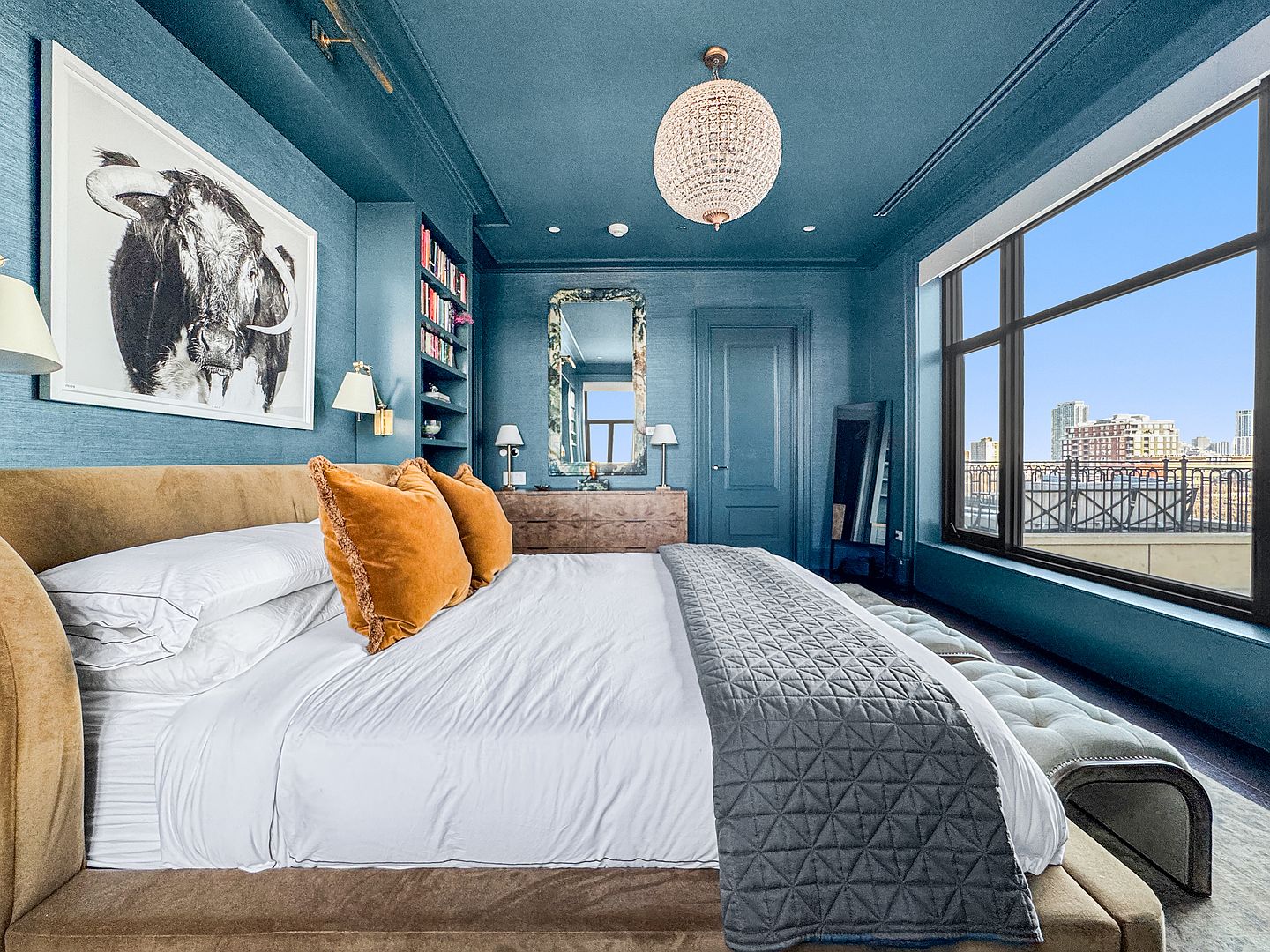 This sophisticated primary bedroom features deep blue walls and a matching ceiling, creating a moody and immersive atmosphere. The room is anchored by a large bed with a tan upholstered headboard, accented by vibrant orange velvet pillows and a textured gray quilt. Large windows provide an expansive view of the city skyline, while a crystal chandelier and built-in shelving add a touch of luxury and functional elegance.