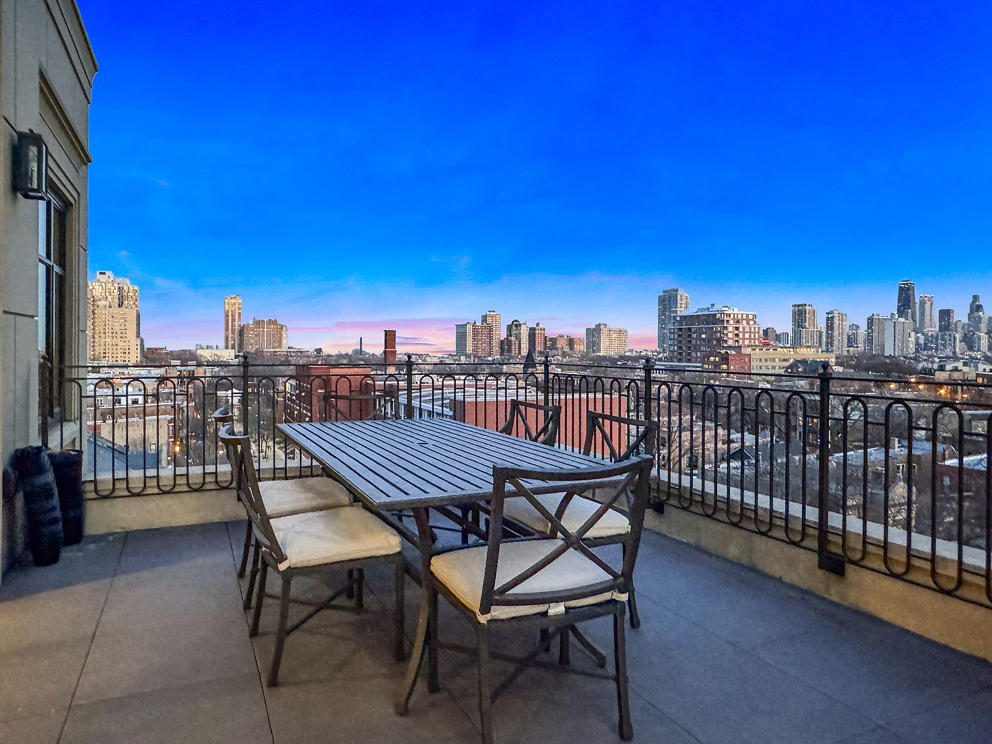 This elevated balcony offers a sophisticated outdoor dining space with a metal table and four matching chairs set against a panoramic urban skyline. The scene is captured at twilight, with the deep blue sky contrasting against the warm, glowing city lights in the distance. A decorative wrought-iron railing encloses the tiled terrace, providing a secure and elegant vantage point for enjoying the city views.