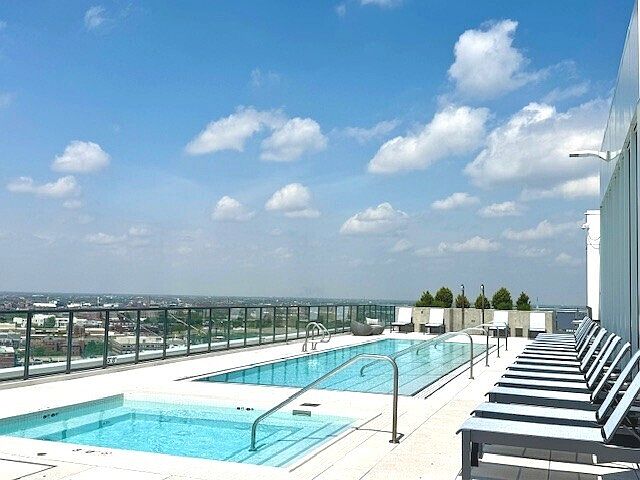 This rooftop amenity space features a rectangular swimming pool and an adjacent square spa, both set against a panoramic city skyline view. A row of modern lounge chairs lines the pool deck, providing a comfortable area for relaxation under the bright blue sky. The perspective is a wide-angle shot from the pool deck, emphasizing the expansive outdoor leisure area and the urban setting.