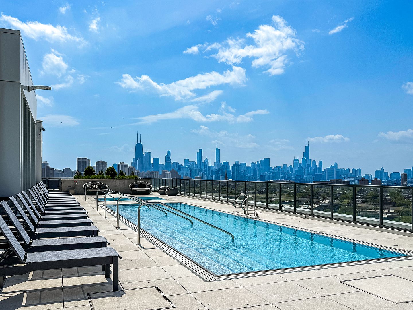 This rooftop pool deck offers a stunning, unobstructed panoramic view of the Chicago skyline. The space features a long, rectangular swimming pool with integrated handrails, surrounded by a clean, paved deck lined with multiple dark-toned lounge chairs. The modern, open-air setting is framed by a sleek metal railing, creating an inviting and sophisticated urban oasis.