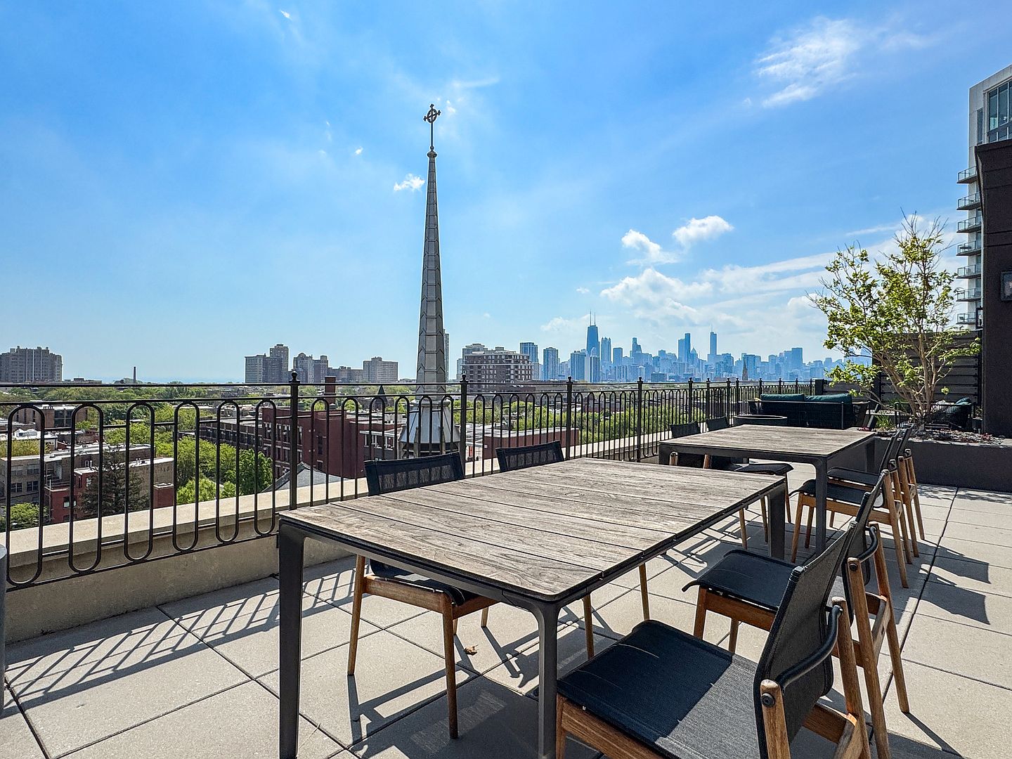 This expansive rooftop terrace offers a stunning panoramic view of the city skyline, featuring a prominent church spire in the foreground. The space is furnished with two large wooden dining tables and matching chairs, providing an ideal setting for outdoor entertaining. The area is enclosed by a black metal railing and paved with large concrete tiles, creating a clean and modern urban retreat.