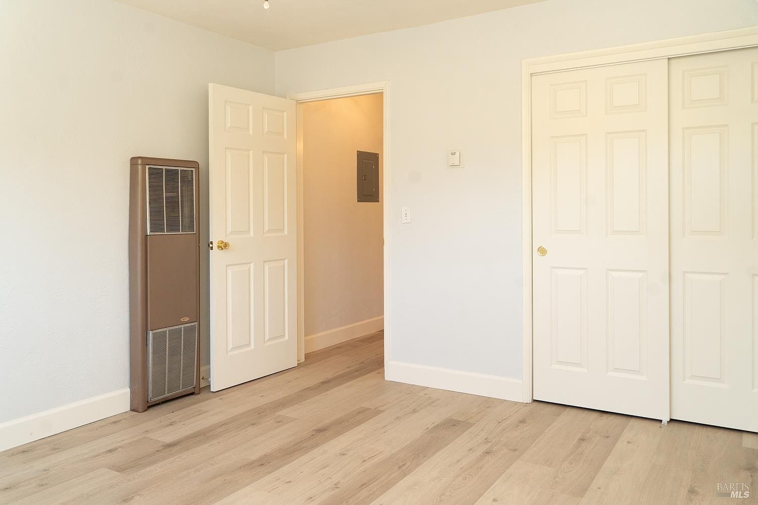 This interior view shows a neutral-toned bedroom featuring light wood-look flooring and white walls. The room includes a wall-mounted heater, a doorway leading to an adjacent space, and a set of white double-panel closet doors. The perspective is straight-on, capturing the simple, clean layout of the room.