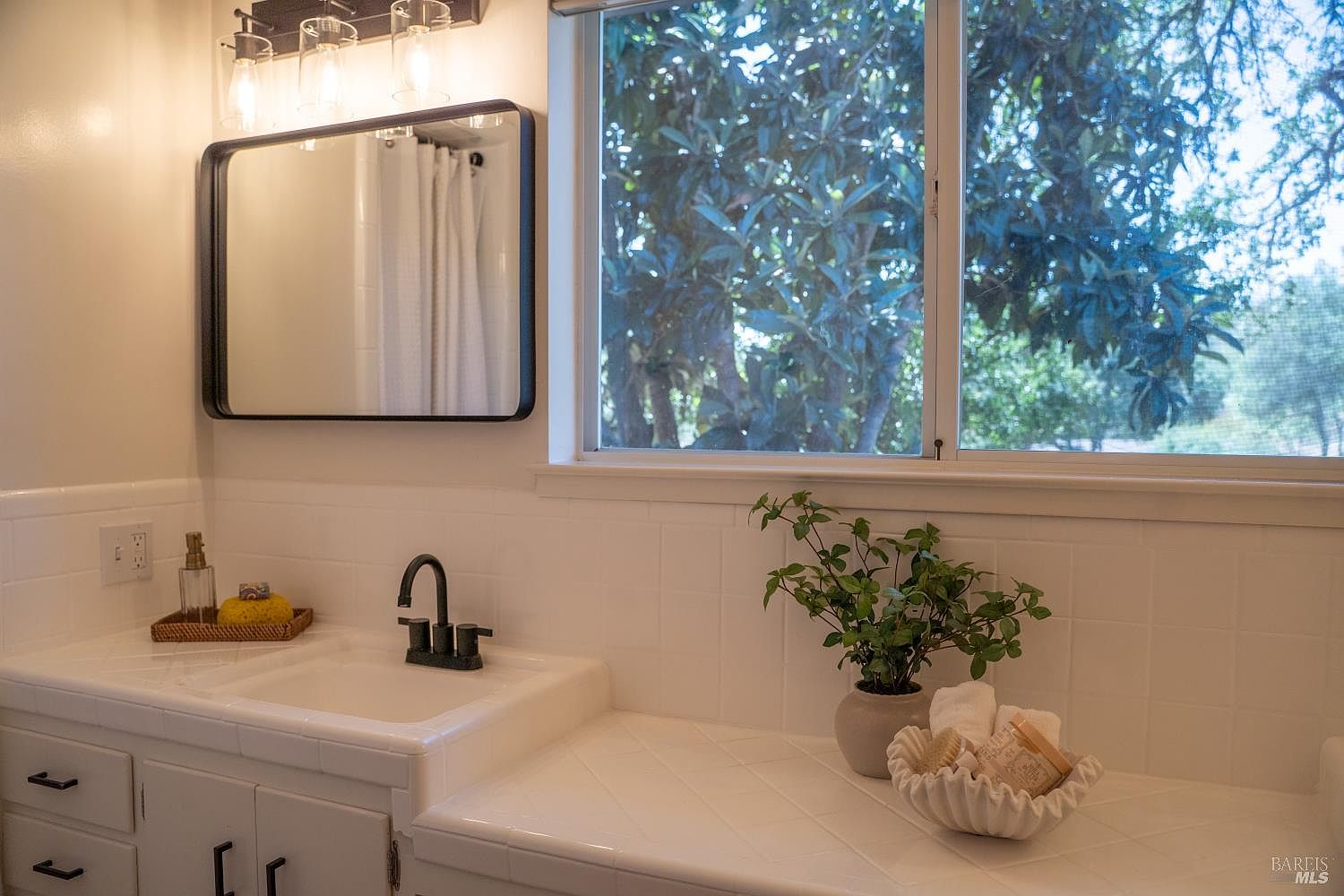 This bright bathroom vanity area features a white tiled countertop with a drop-in sink and a matte black faucet. A rectangular mirror with a dark frame hangs above the sink, complemented by a three-light fixture, while a large window provides natural light and a view of the outdoors. The space is accented with a potted plant and a decorative bowl containing towels, creating a clean and inviting atmosphere.