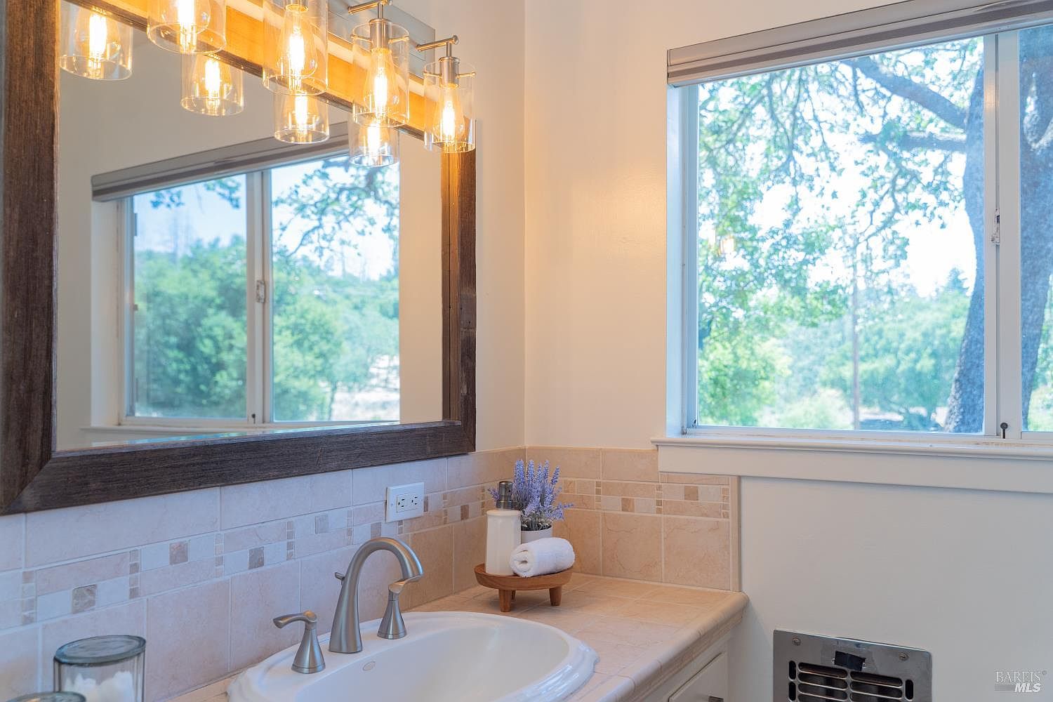 This bathroom features a vanity with a light-toned tile countertop and a white undermount sink, complemented by a brushed-nickel faucet. A large mirror with a dark wood frame hangs above the vanity, reflecting the window and the light fixture with multiple glass shades. The walls are finished with neutral-toned tiles, and a small decorative arrangement of lavender sits on the counter, creating a clean and inviting atmosphere.