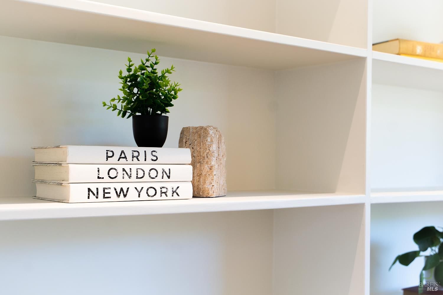This close-up shot highlights a section of clean, white built-in shelving, showcasing a minimalist and organized aesthetic. The shelves are styled with a small potted plant, a textured stone accent piece, and a stack of decorative books labeled with city names. The bright, neutral palette creates a crisp and inviting atmosphere suitable for a modern home interior.
